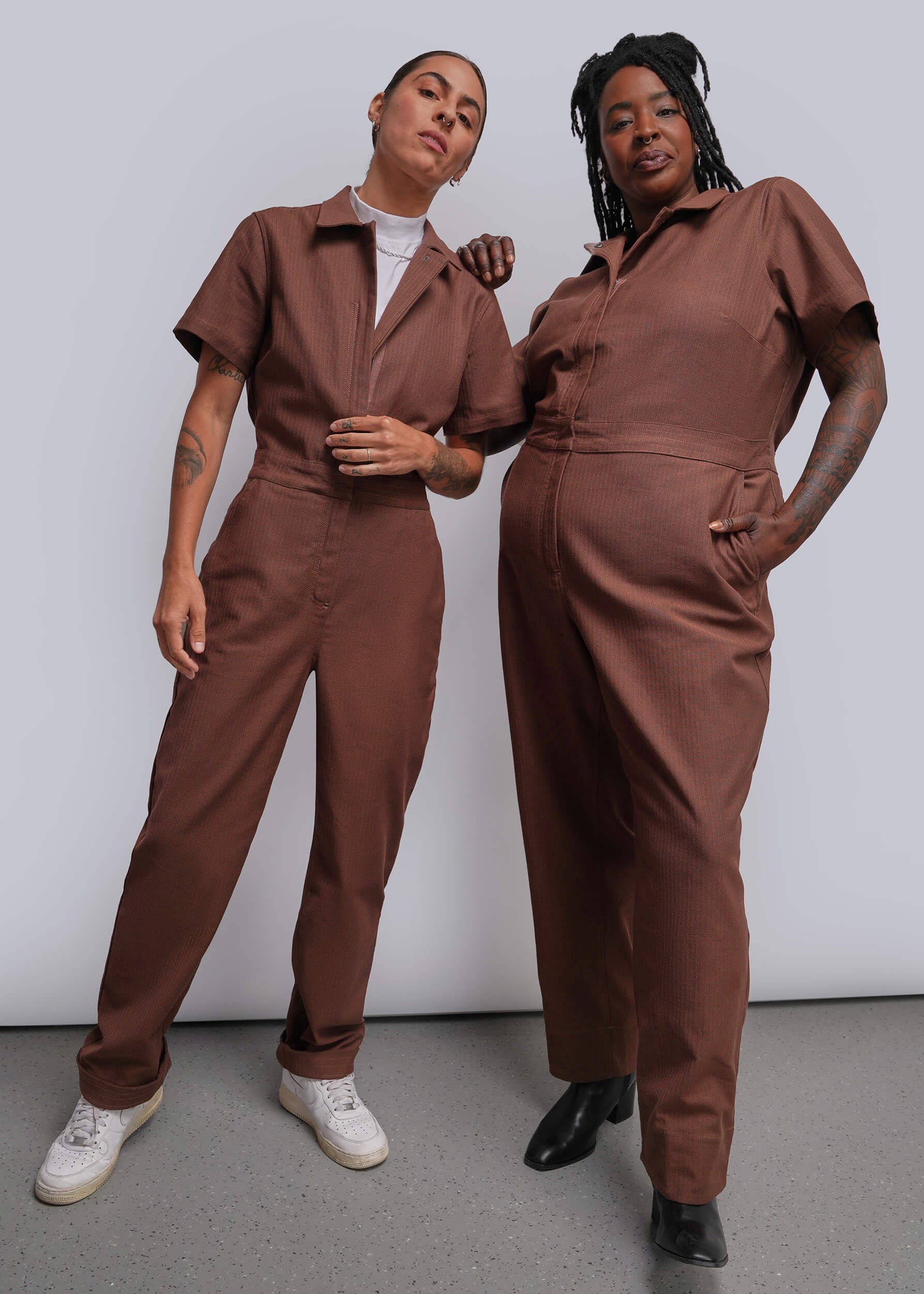 Two models wearing matching brown short-sleeve utility jumpsuits, standing against a plain light gray studio background.