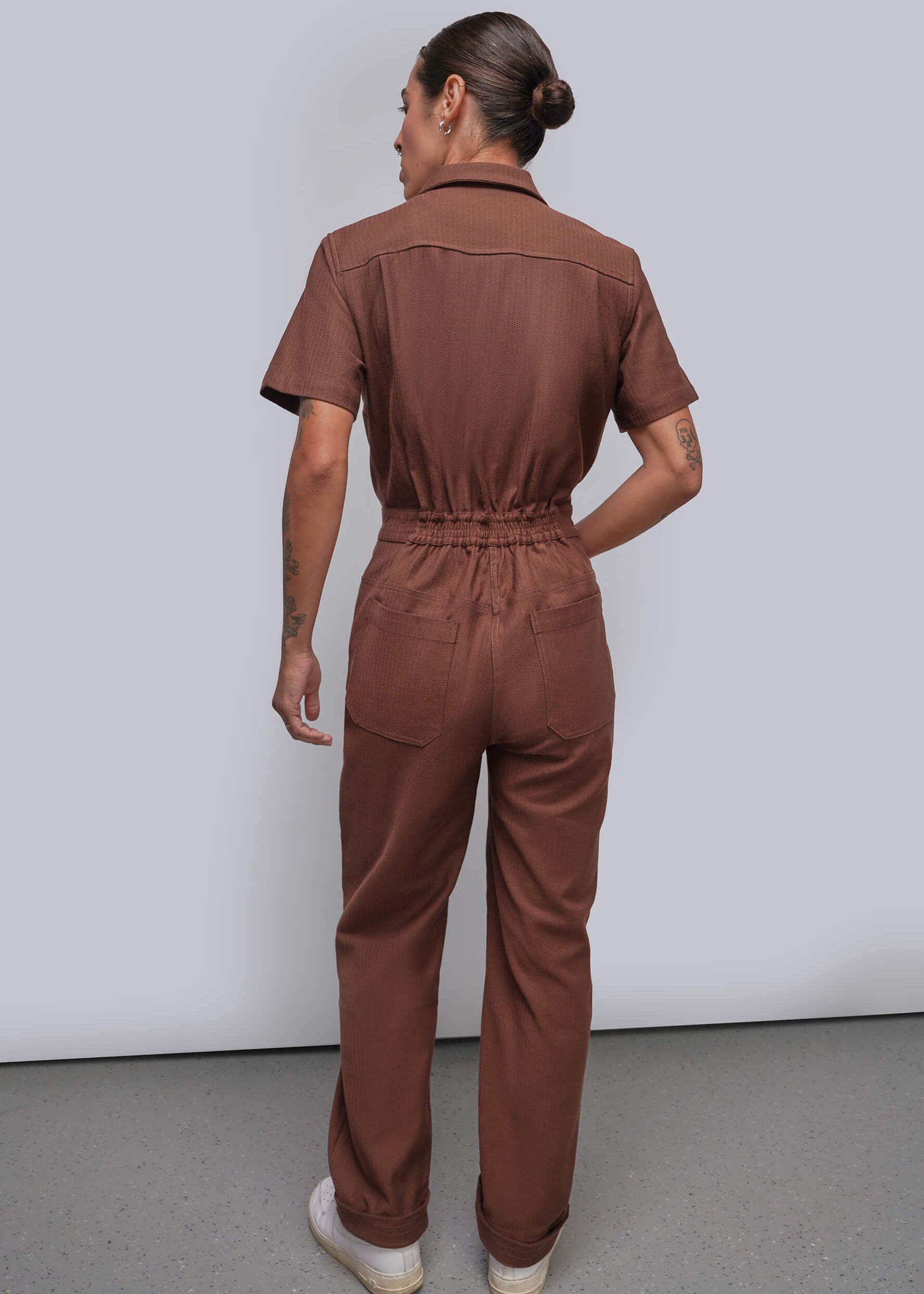 S=A model shown from the back wearing a brown short-sleeve jumpsuit with a gathered elastic waist, set against a plain light gray studio background.