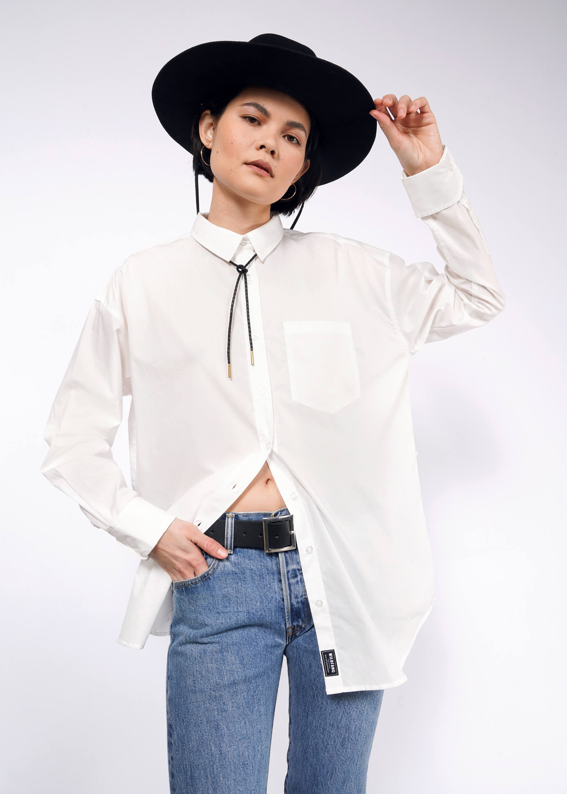 S= Person posing against a white background wearing The Essential Long Sleeve Oversized Wrap Button Up with French cuffs, bolo tie, blue jeans, and a wide-brimmed black hat. The shirt is partially tucked in, and they hold the brim of the hat with one hand.
