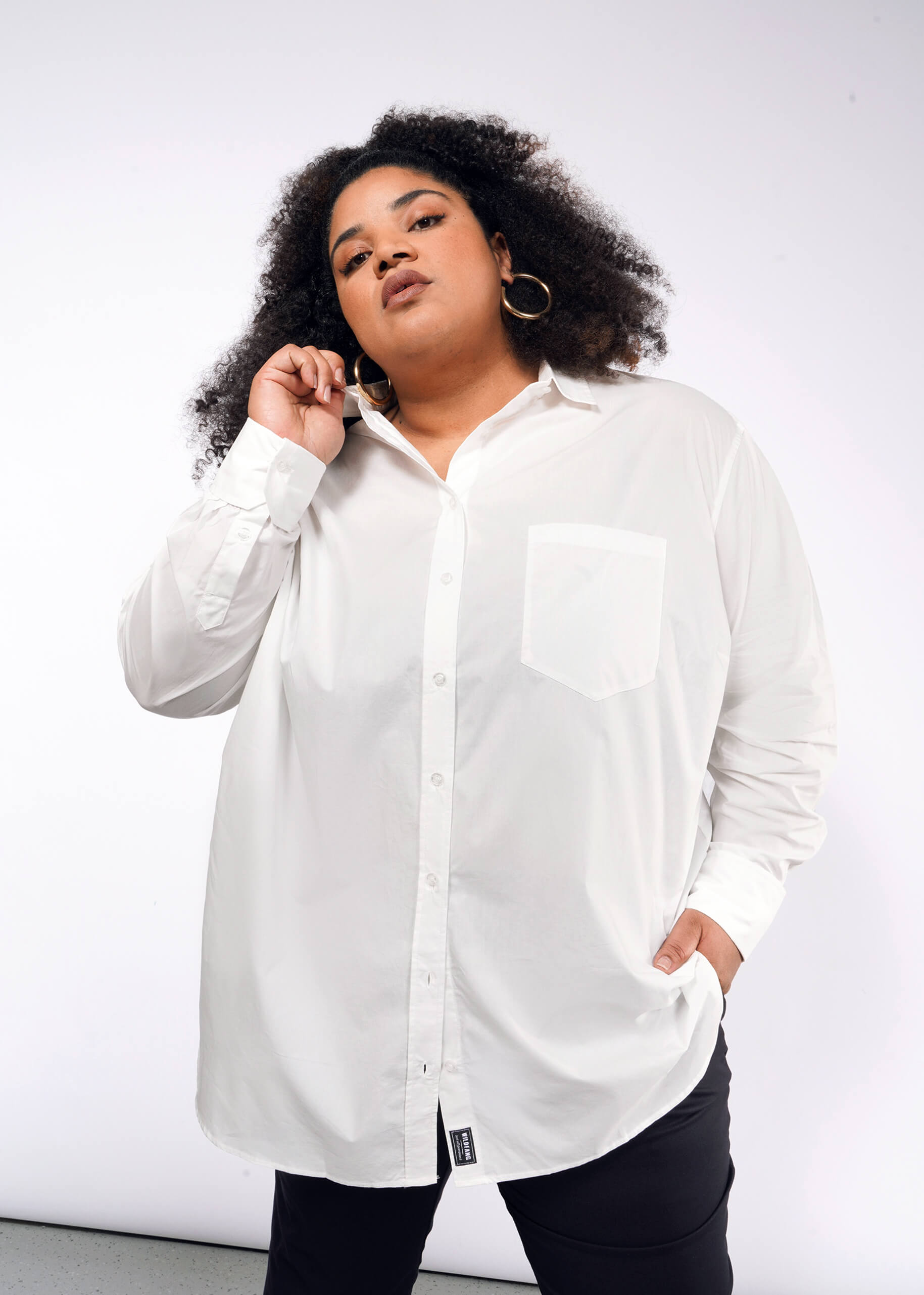 1X= A person with curly hair and large hoop earrings poses confidently, wearing The Essential Long Sleeve Oversized Wrap Button Up shirt with French cuffs and black pants. One hand touches the collar while the other rests in a pocket against a plain white background.