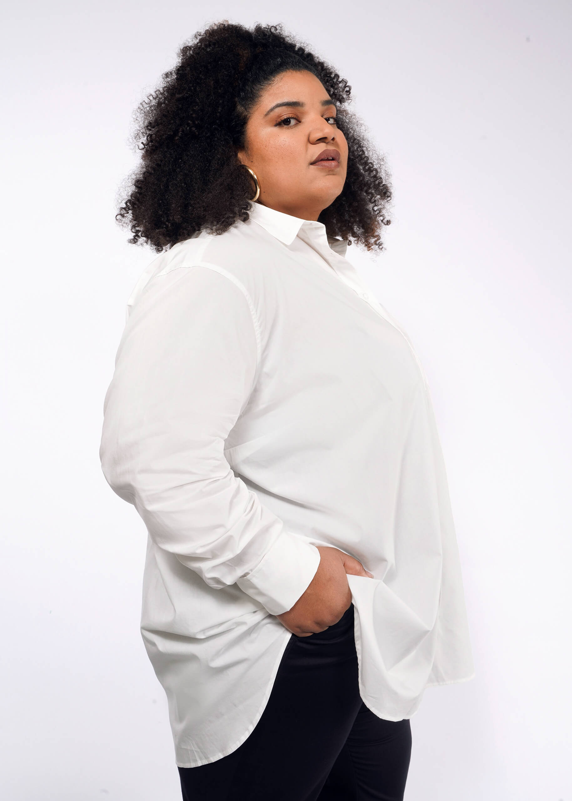 1X=A person with curly hair confidently stands against a plain background, wearing The Essential Long Sleeve Oversized Wrap Button Up shirt and black pants, with one hand in their pocket.