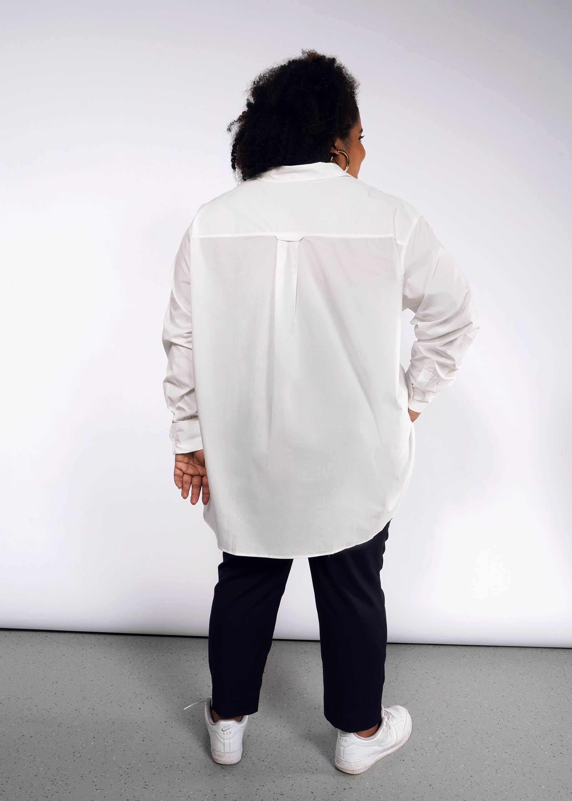 1X= A person with curly hair, facing away from the camera, wears The Essential Long Sleeve Oversized Wrap Button Up with French cuffs, black pants, and white sneakers. A light gray wall and floor create a minimalist backdrop.
