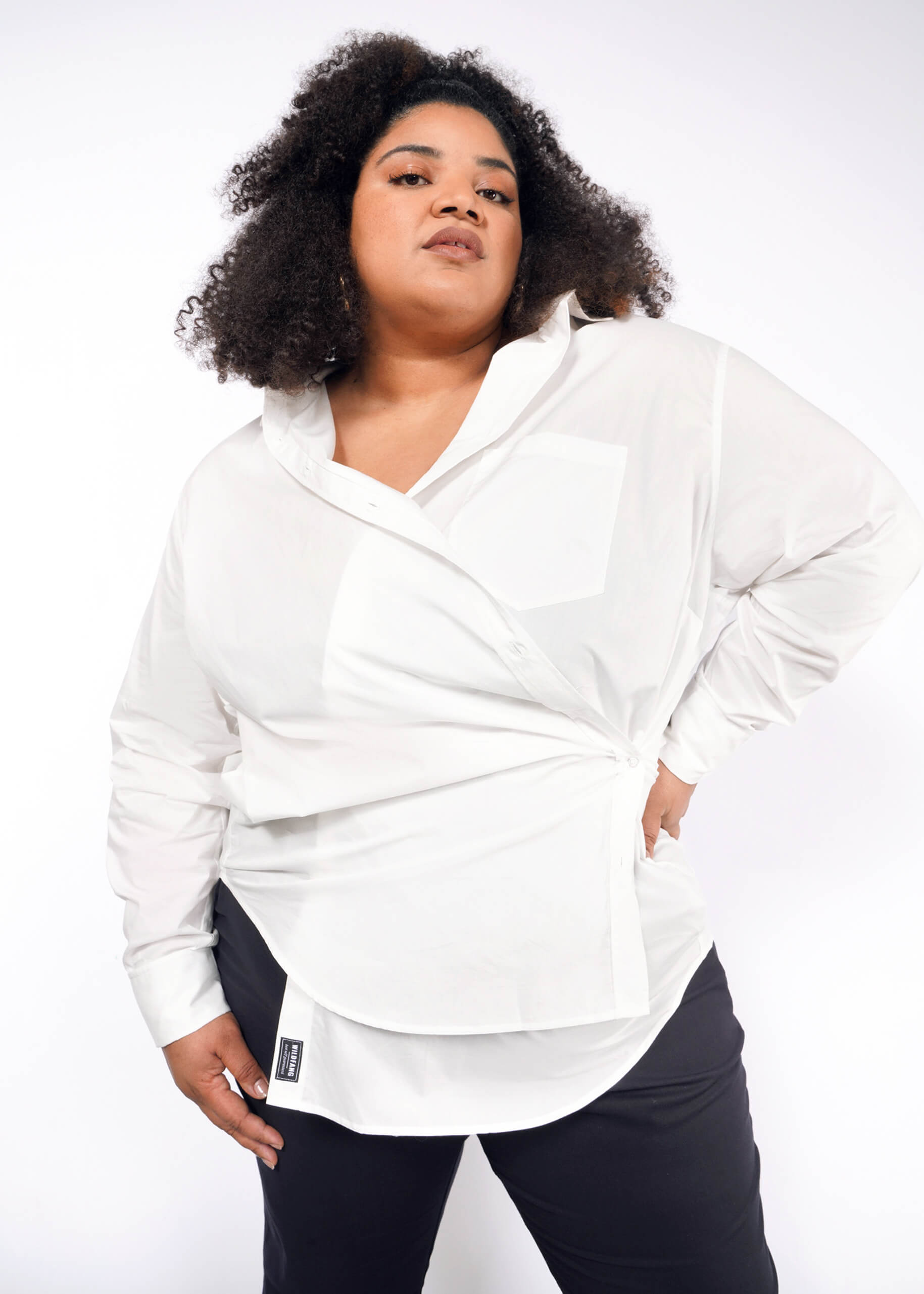 1X=A person with voluminous curly hair confidently poses in The Essential Long Sleeve Oversized Wrap Button Up and black pants, one hand in a pocket and the other by their side against a plain white background.