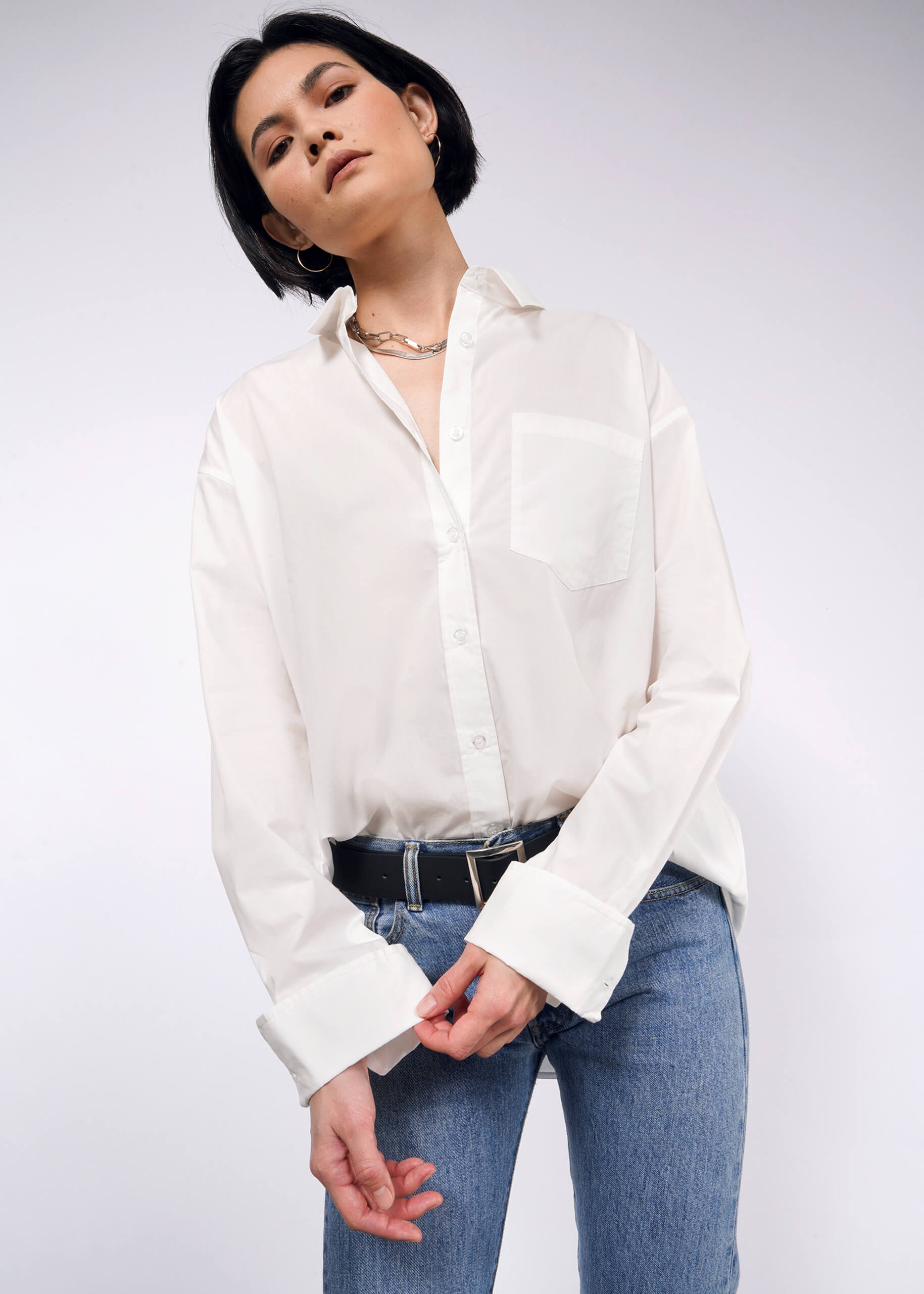 S=A person with short dark hair is wearing The Essential Long Sleeve Oversized Wrap Button Up in white, paired with blue jeans and a black belt. They pose against a plain background, gazing slightly downward.