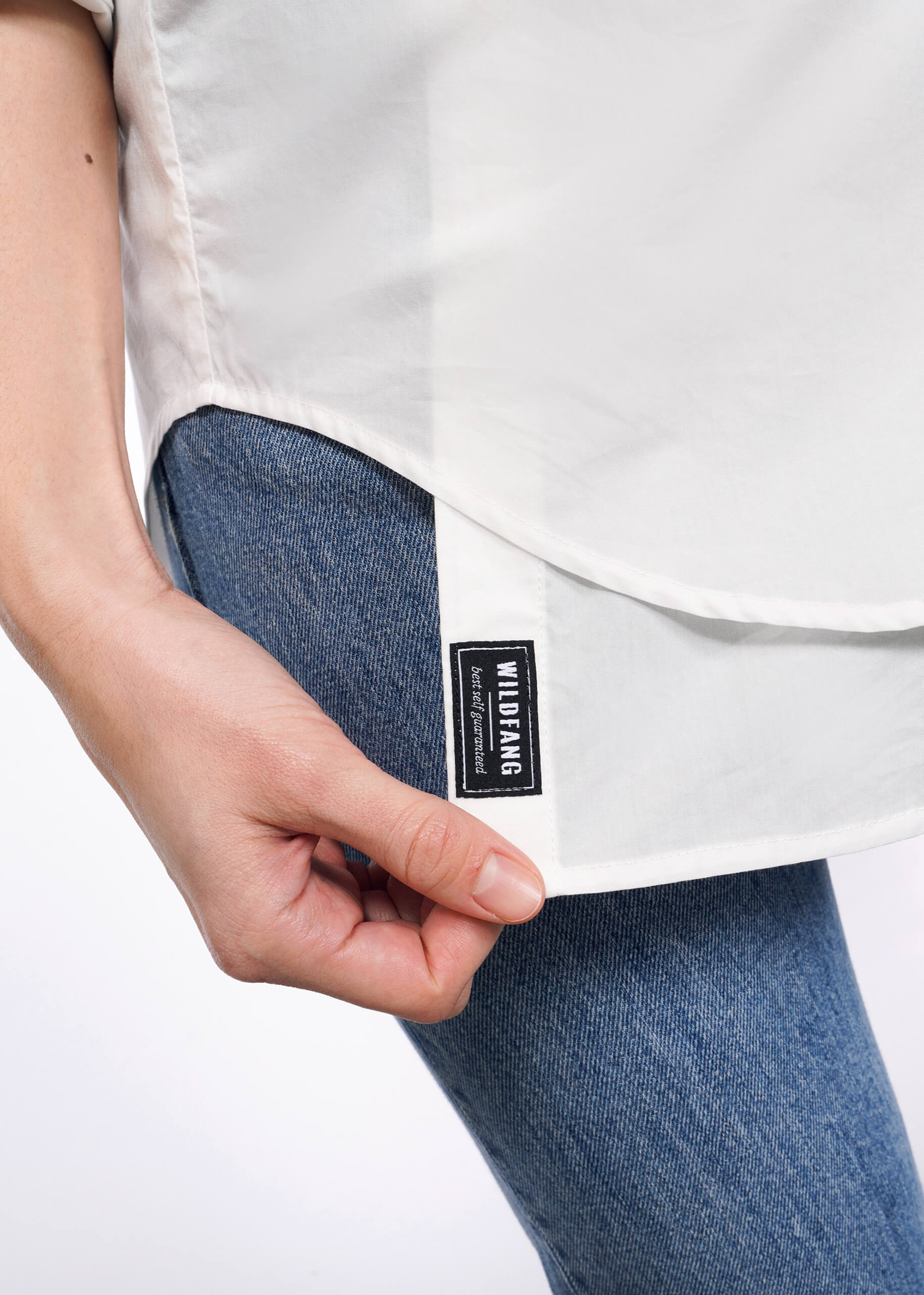 S=Close-up of a person holding the edge of The Essential Long Sleeve Oversized Wrap Button Up in white, featuring a black Wu-Tang Brand Limited tag. They are wearing blue jeans, with focus on the shirt and tag against a plain white background.