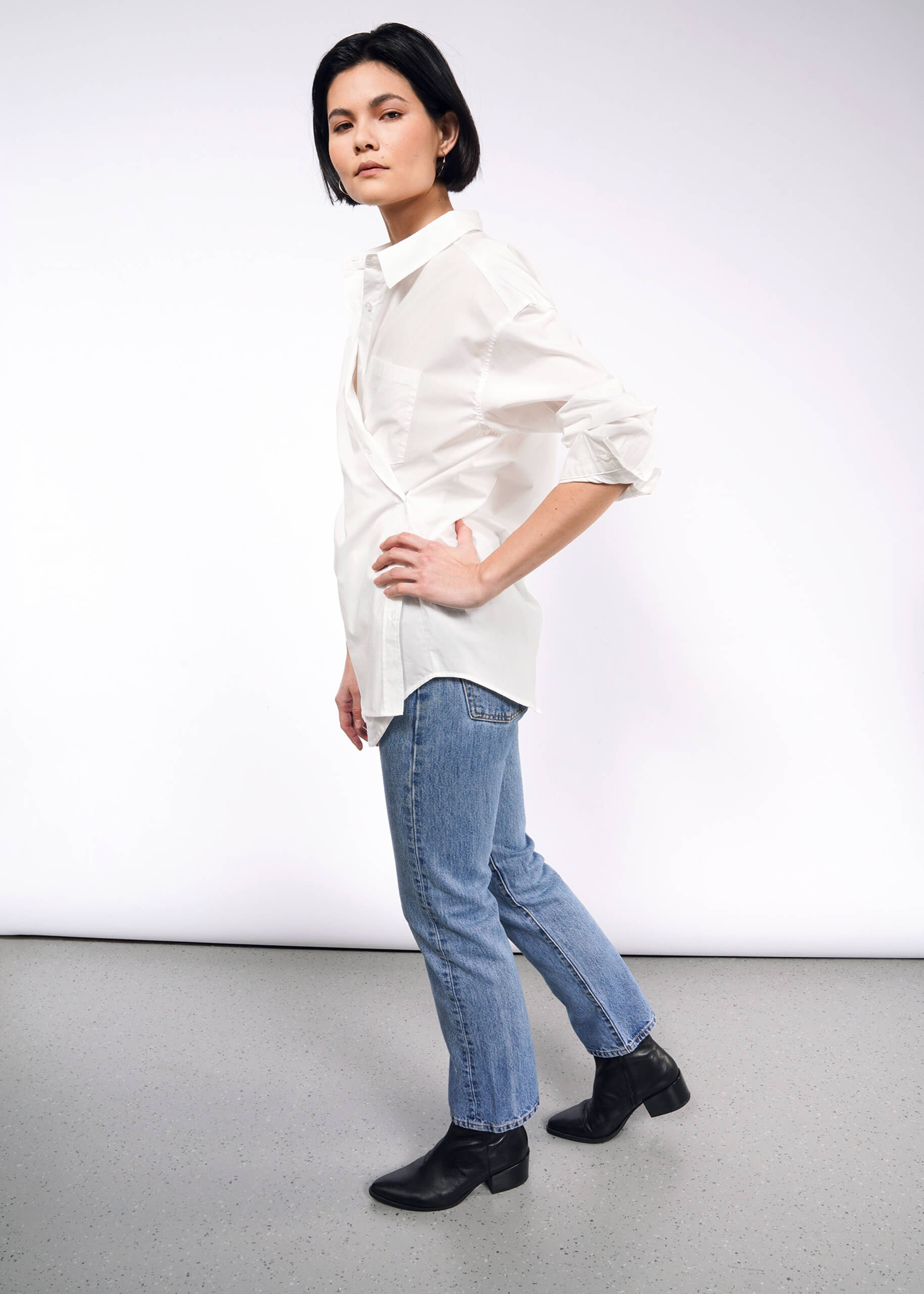S=Against a plain white background, a person with short dark hair stands confidently, wearing The Essential Long Sleeve Oversized Wrap Button Up shirt with blue jeans. They pair this look with black ankle boots and have one hand on their hip, enhancing the outfits sophistication with French cuffs.