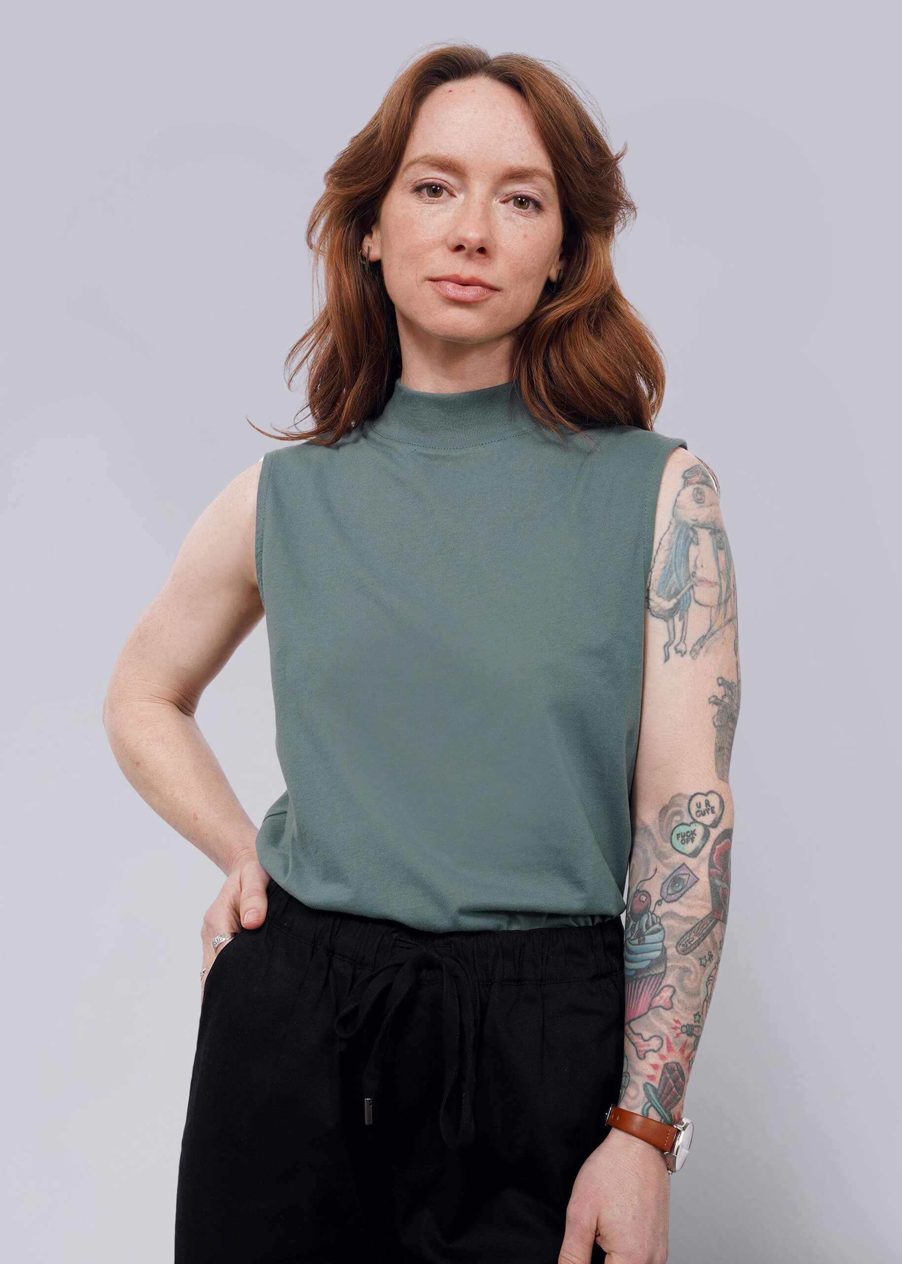 S= A woman with long brown hair and visible tattoos on her left arm stands confidently against a plain white background, wearing The Essential Mock Neck Tank in green paired with black pants.