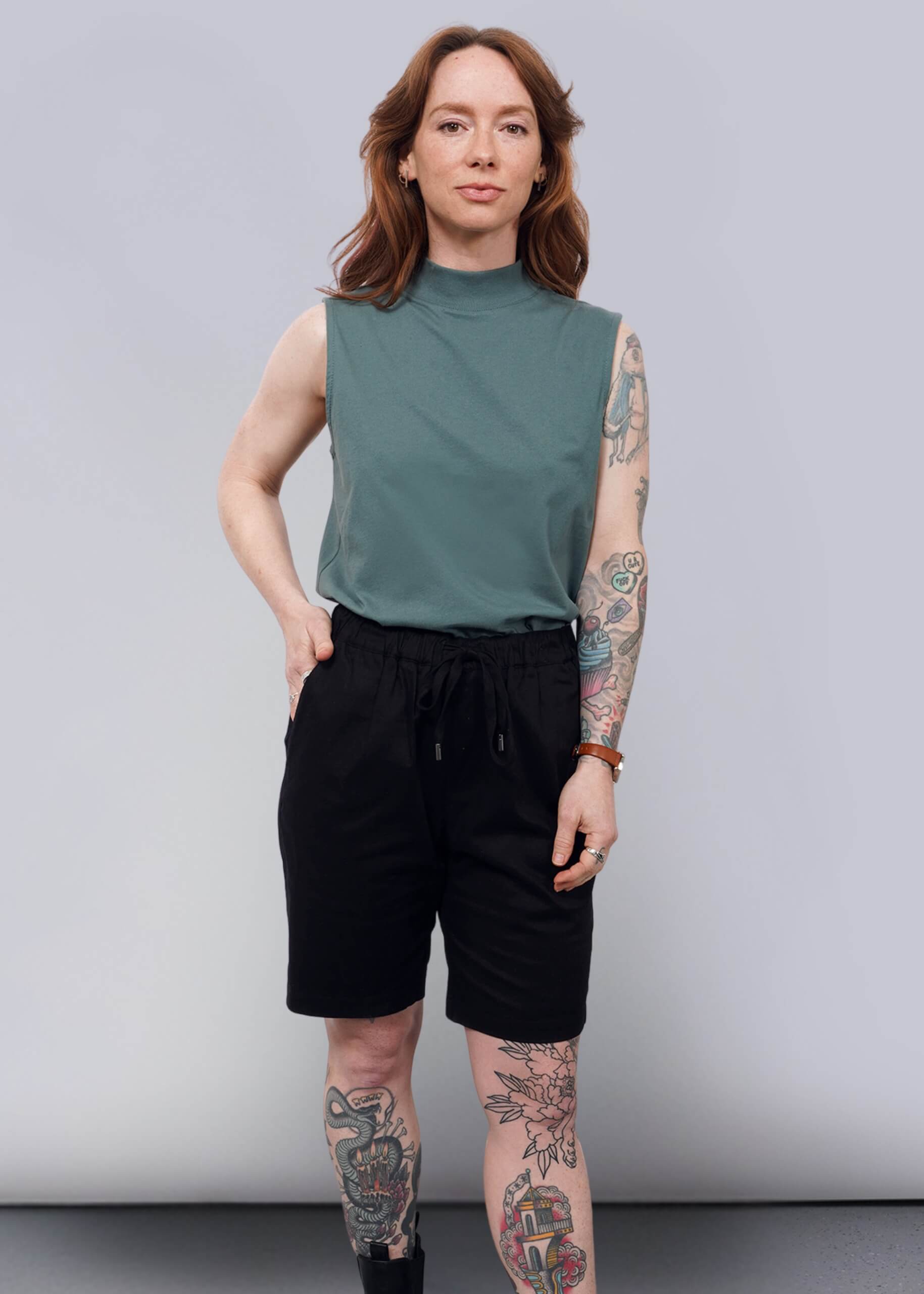 S=A person with long, wavy brown hair and visible tattoos stands confidently against a plain white wall, styled in The Essential Mock Neck Tank in green with black shorts and boots—a perfect layering piece for any casual outfit.