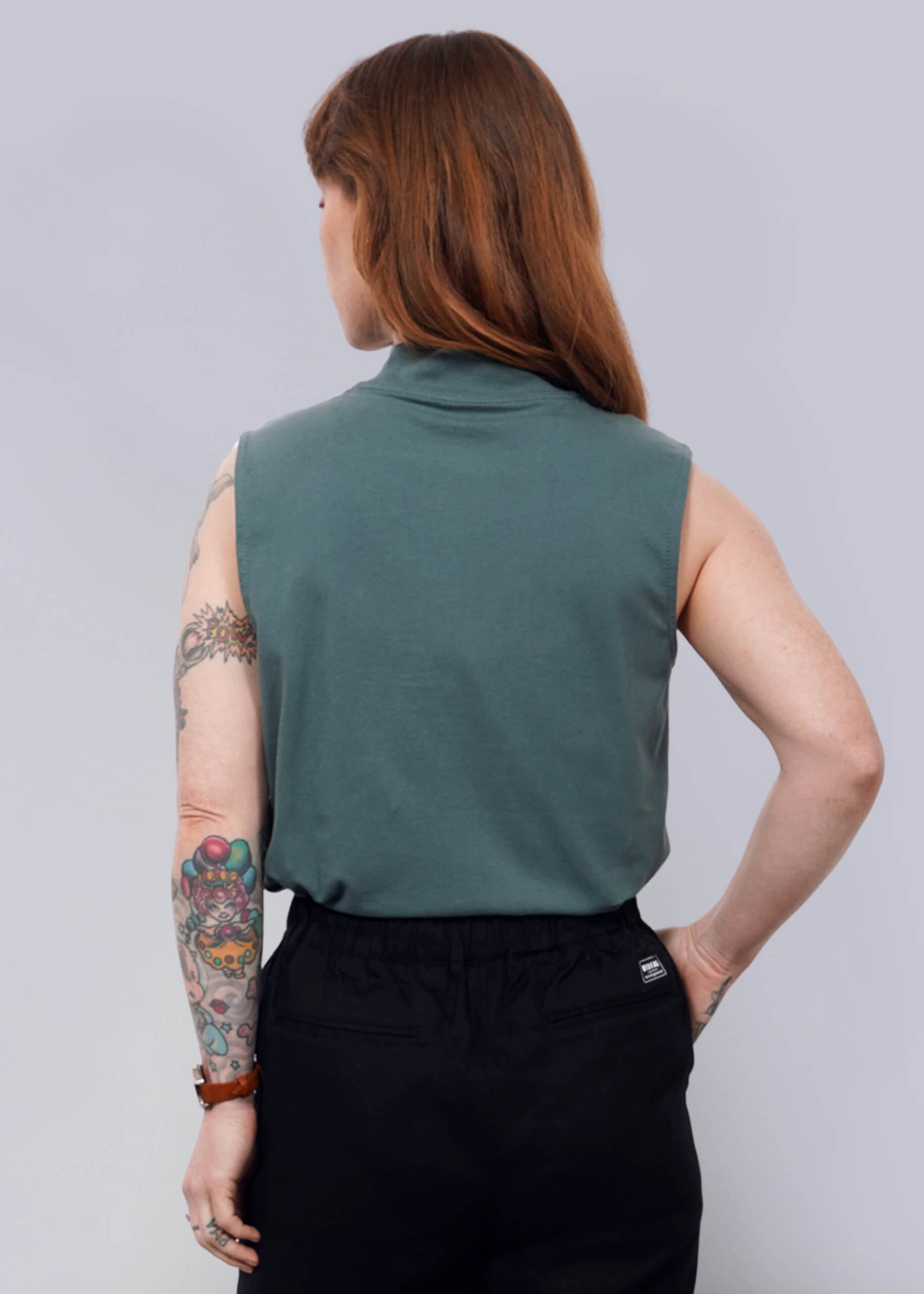 S=A person with long hair and visible tattoos wears The Essential Mock Neck Tank in green, paired with black shorts and black shoes, standing with their back to the camera against a plain white background.