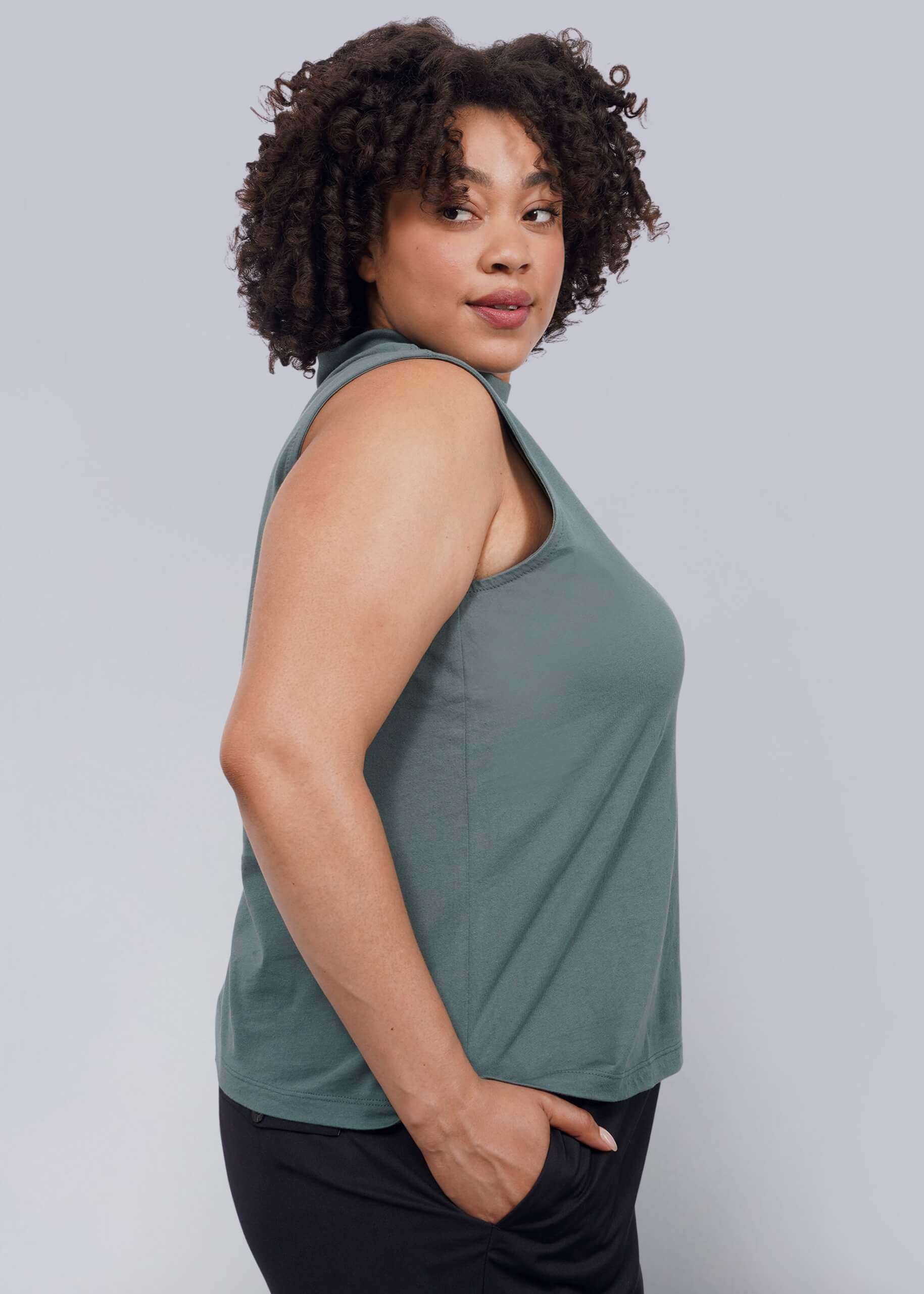 1X= A woman with curly hair wears The Essential Mock Neck Tank in green and black pants, standing in profile against a white background and looking over her shoulder with a slight smile.
