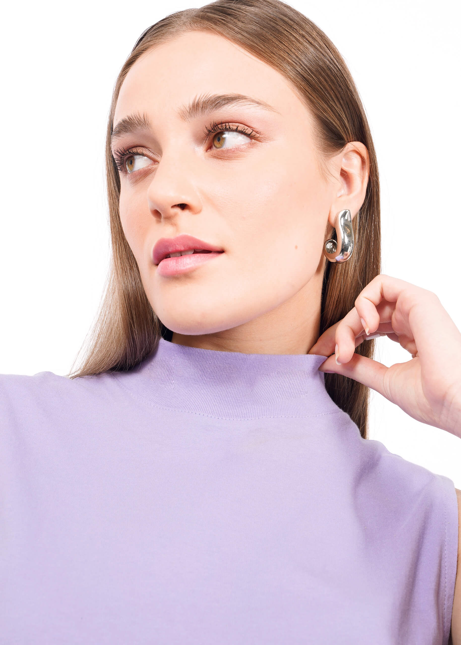 A woman with straight brown hair wears The Essential Mock Neck Tank in lavender—a relaxed-fit, sleeveless layering piece—paired with large silver earrings. She gazes calmly to the side, holding the tops collar with one hand.