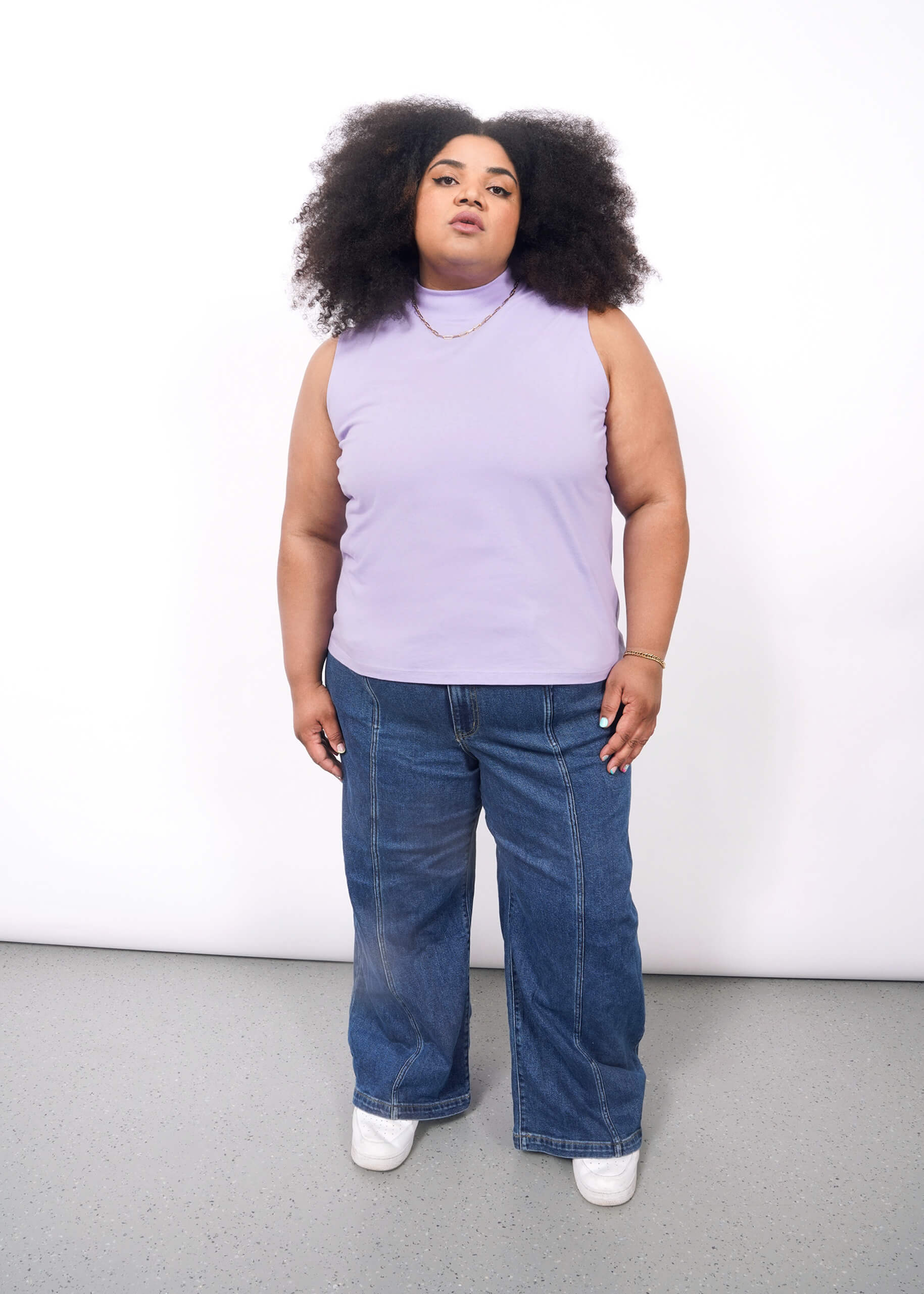 1X=A person with voluminous curly hair wears The Essential Mock Neck Tank in lavender, paired with wide-leg blue jeans, white sneakers, and gold jewelry, standing against a plain white background.