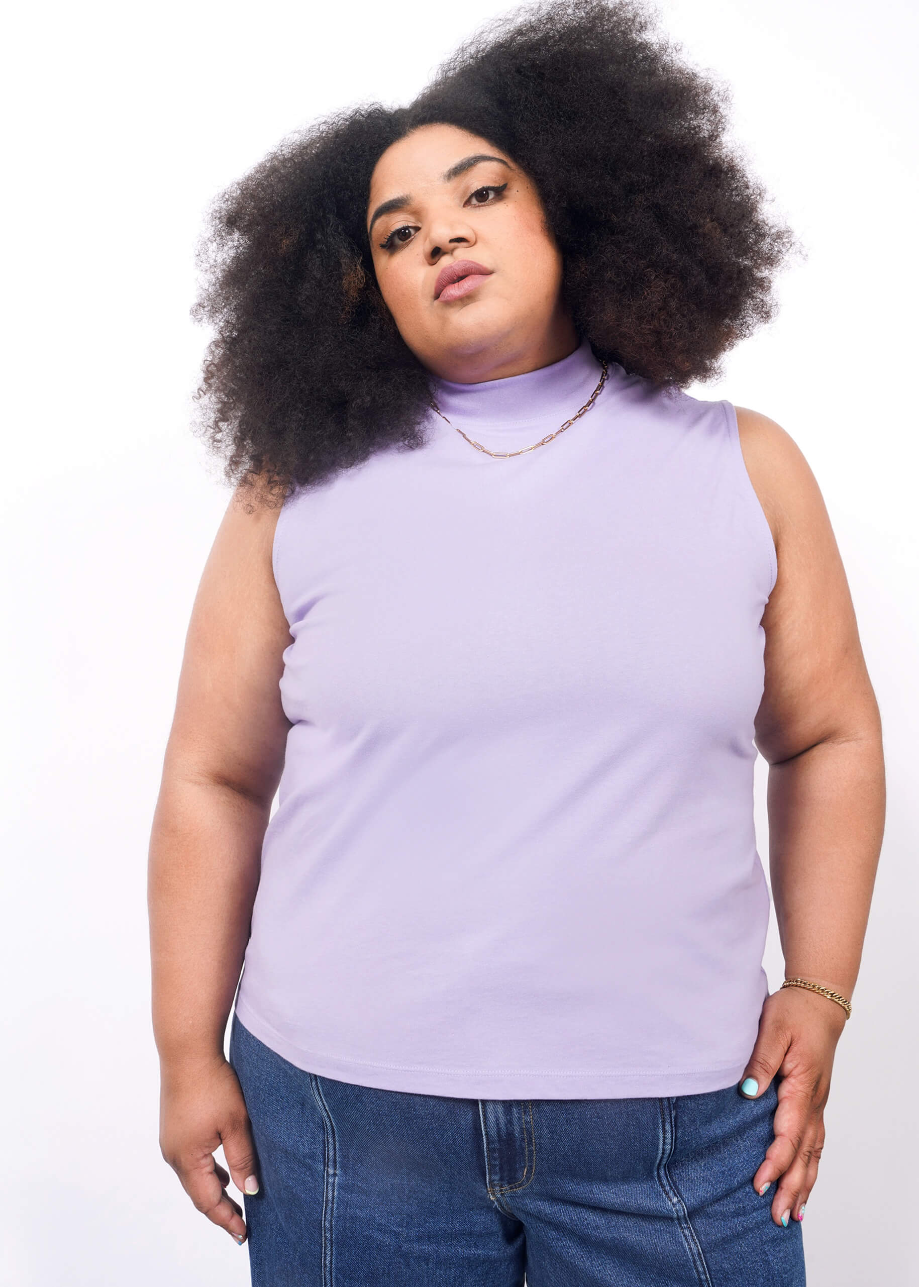 1X= A person with voluminous curly hair wears The Essential Mock Neck Tank in lavender and denim pants, accessorized with a gold chain necklace and bracelet, standing confidently against a white background.