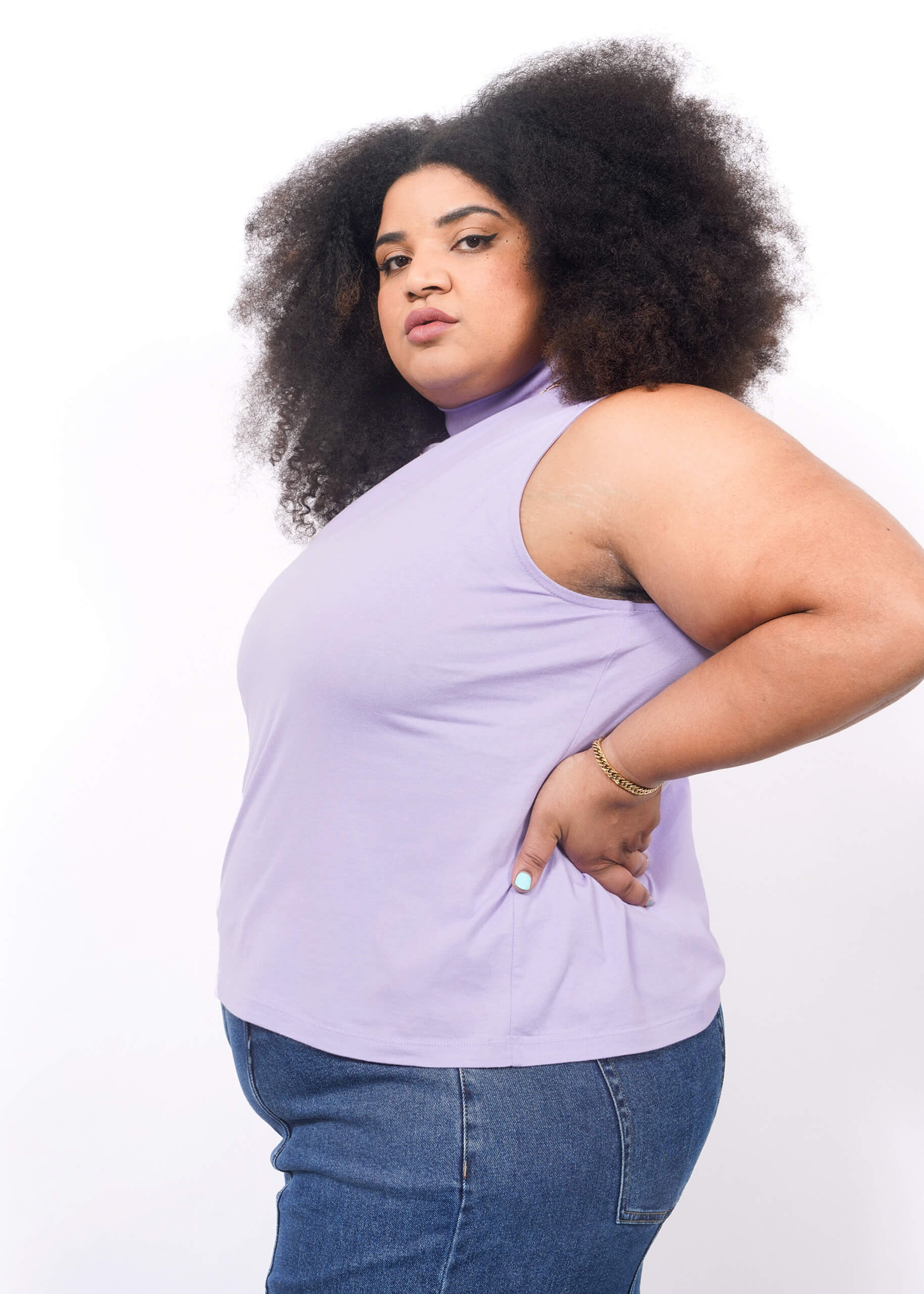 1X= A woman with voluminous curly hair wears The Essential Mock Neck Tank in lavender and blue jeans. Standing in profile against a white background, she looks confidently at the camera with one hand on her hip.