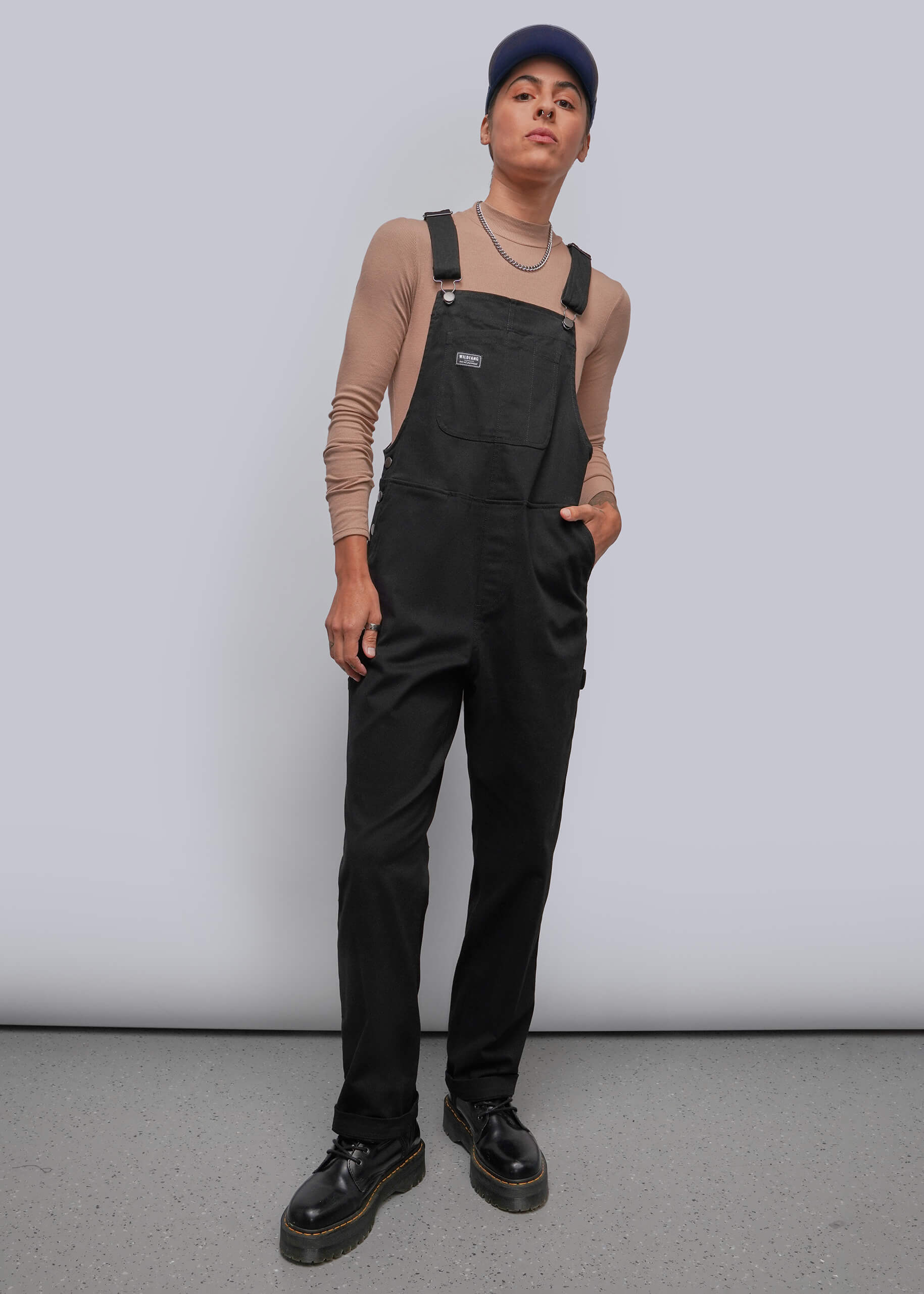 S=A model wearing black overalls over a beige long-sleeve top, paired with a navy blue cap, layered necklaces, and black leather shoes, standing against a light gray studio background.