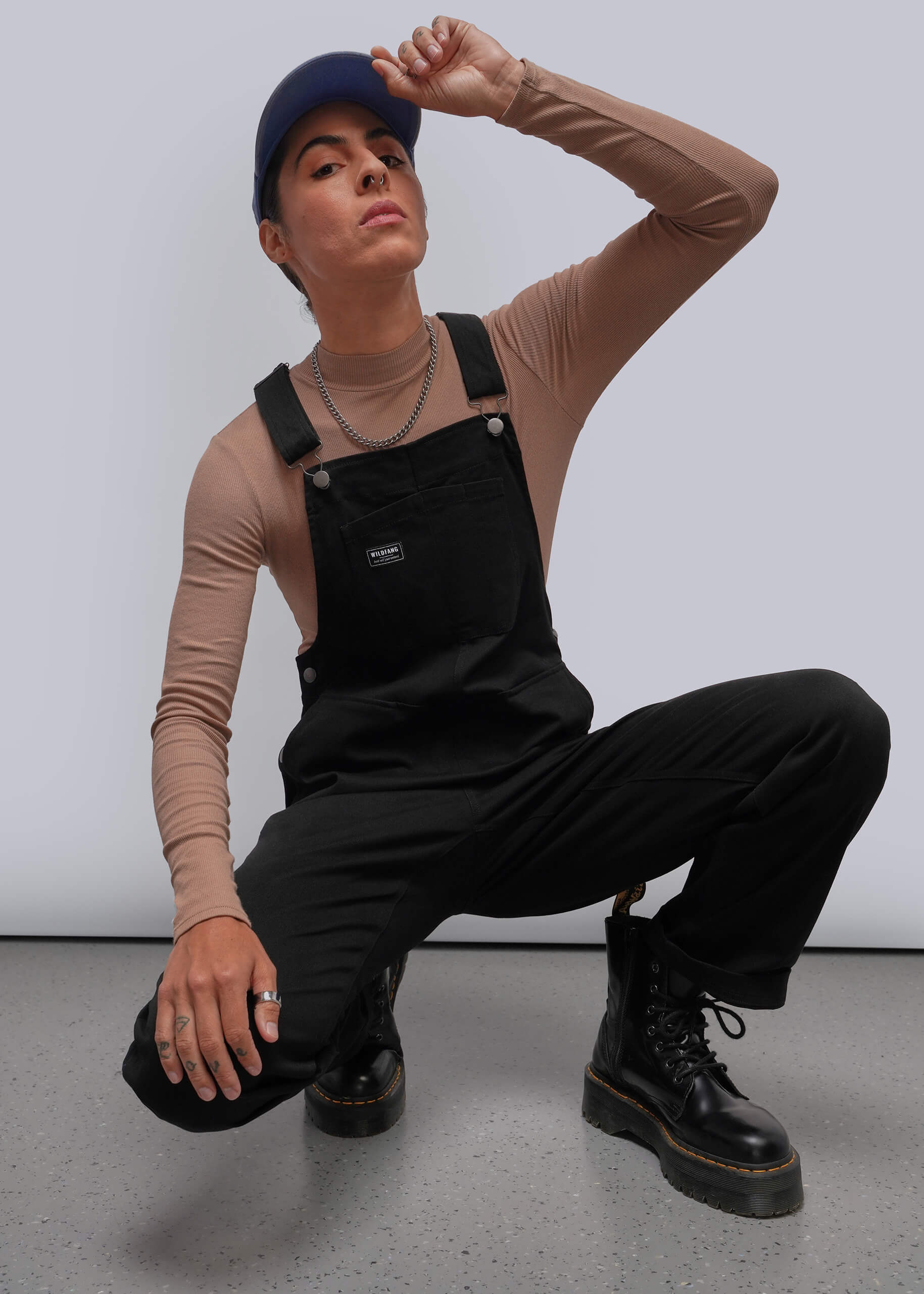 S=A model crouches in a studio wearing black overall-style bib pants over a beige long-sleeve top, with black boots, a dark cap, and silver chain necklaces against a light gray background.