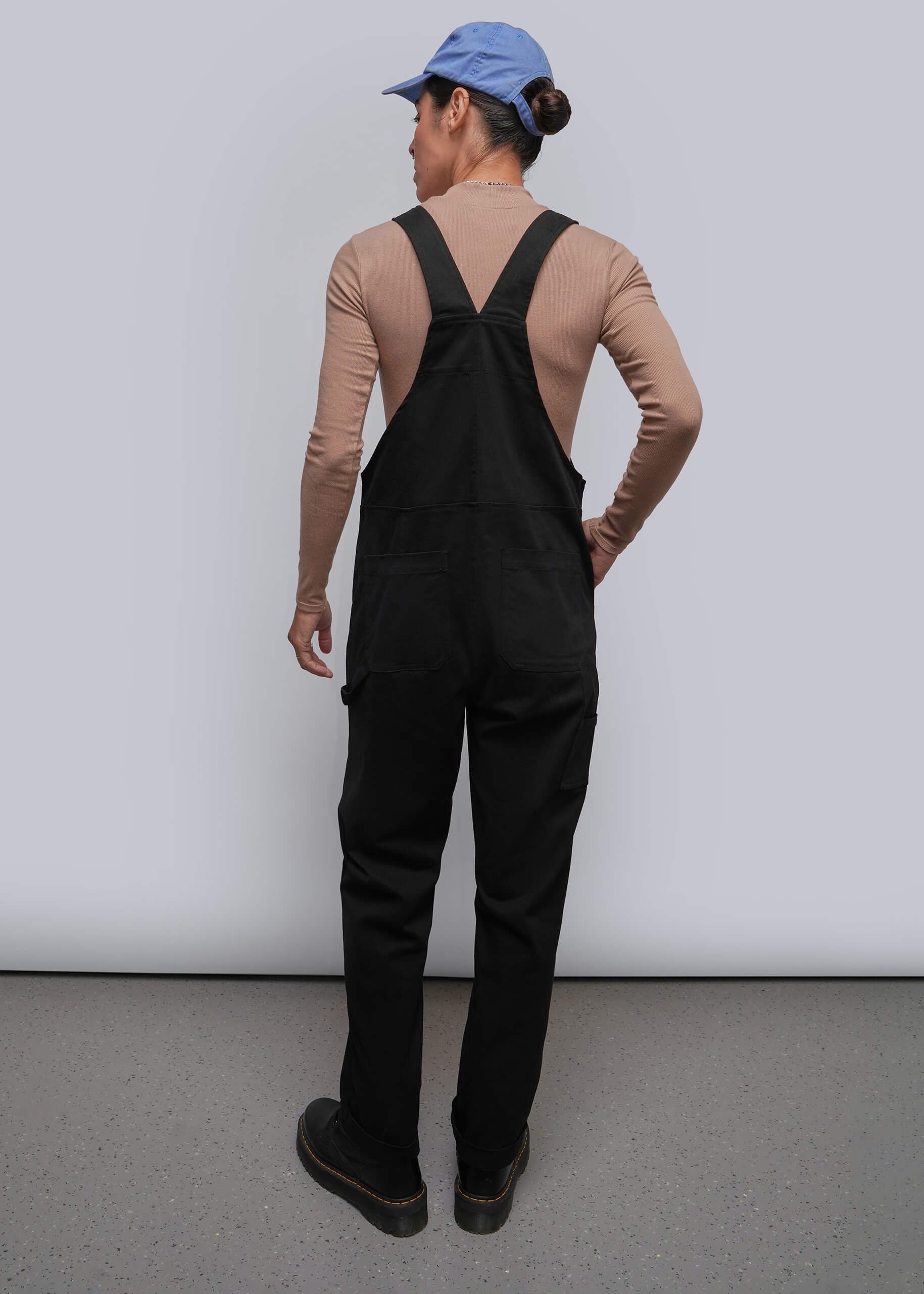 S=A person shown from the back wearing black overalls over a beige long-sleeve top, with a blue cap and dark shoes, standing against a plain light gray wall.
