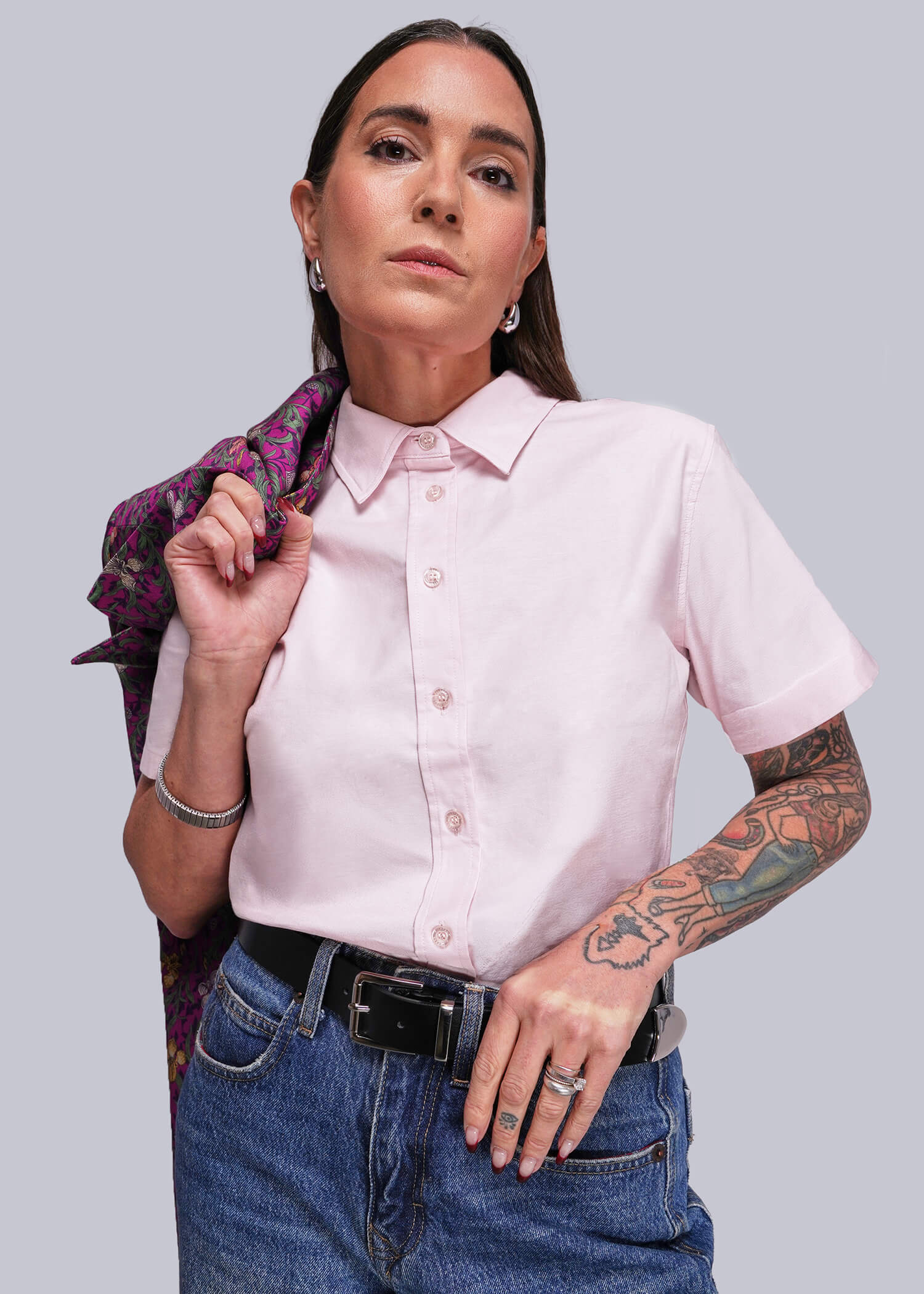 Woman wearing a pink shirt and blue jeans with a patterned blazer over her shoulder, standing against a plain background.