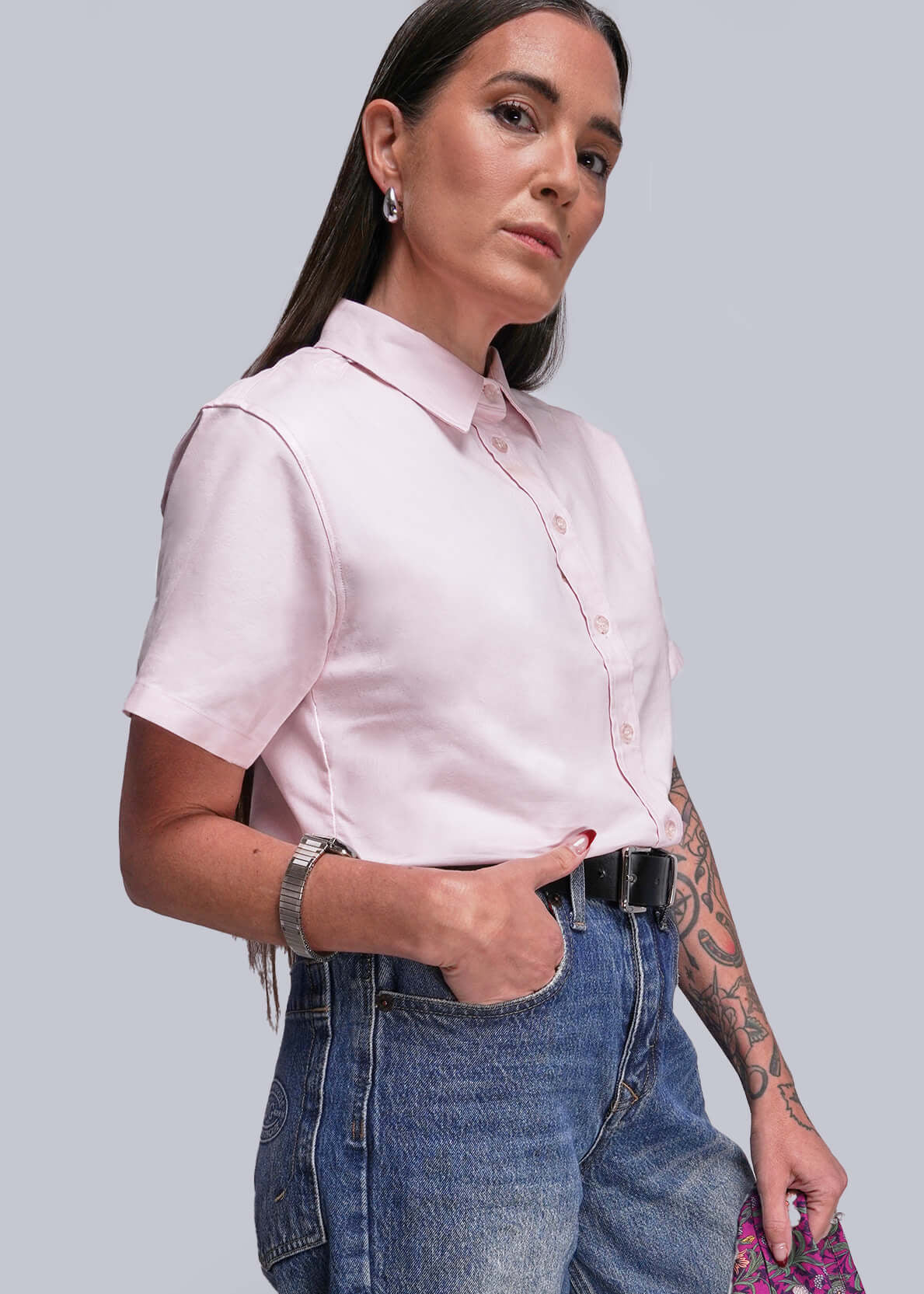 Woman wearing a pink shirt, blue jeans, and a black belt standing against a plain background.