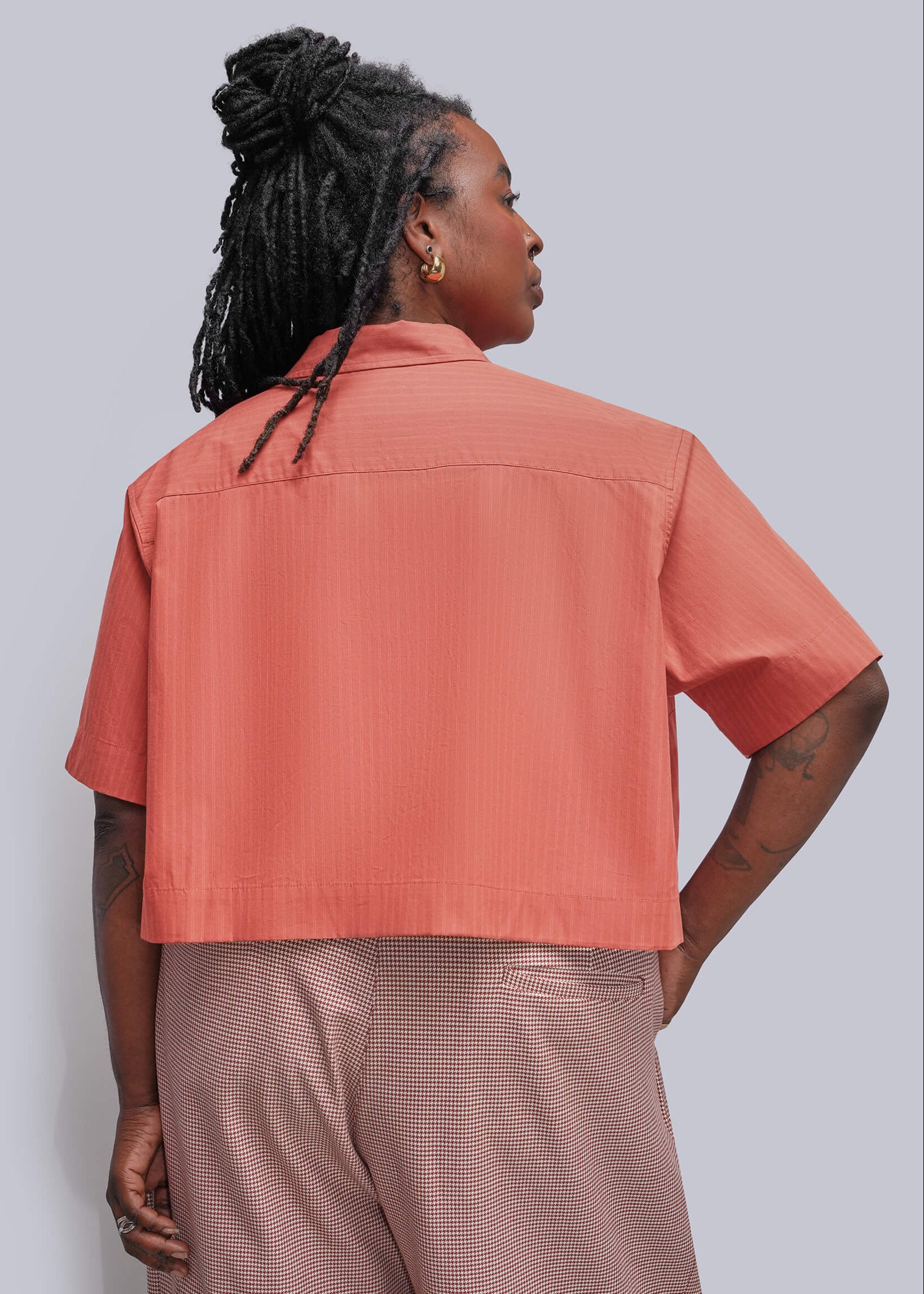 1X=A person shown from the back wearing a short-sleeve collared cropped top in a muted coral and herringbone textured trousers, against a plain light gray studio background.