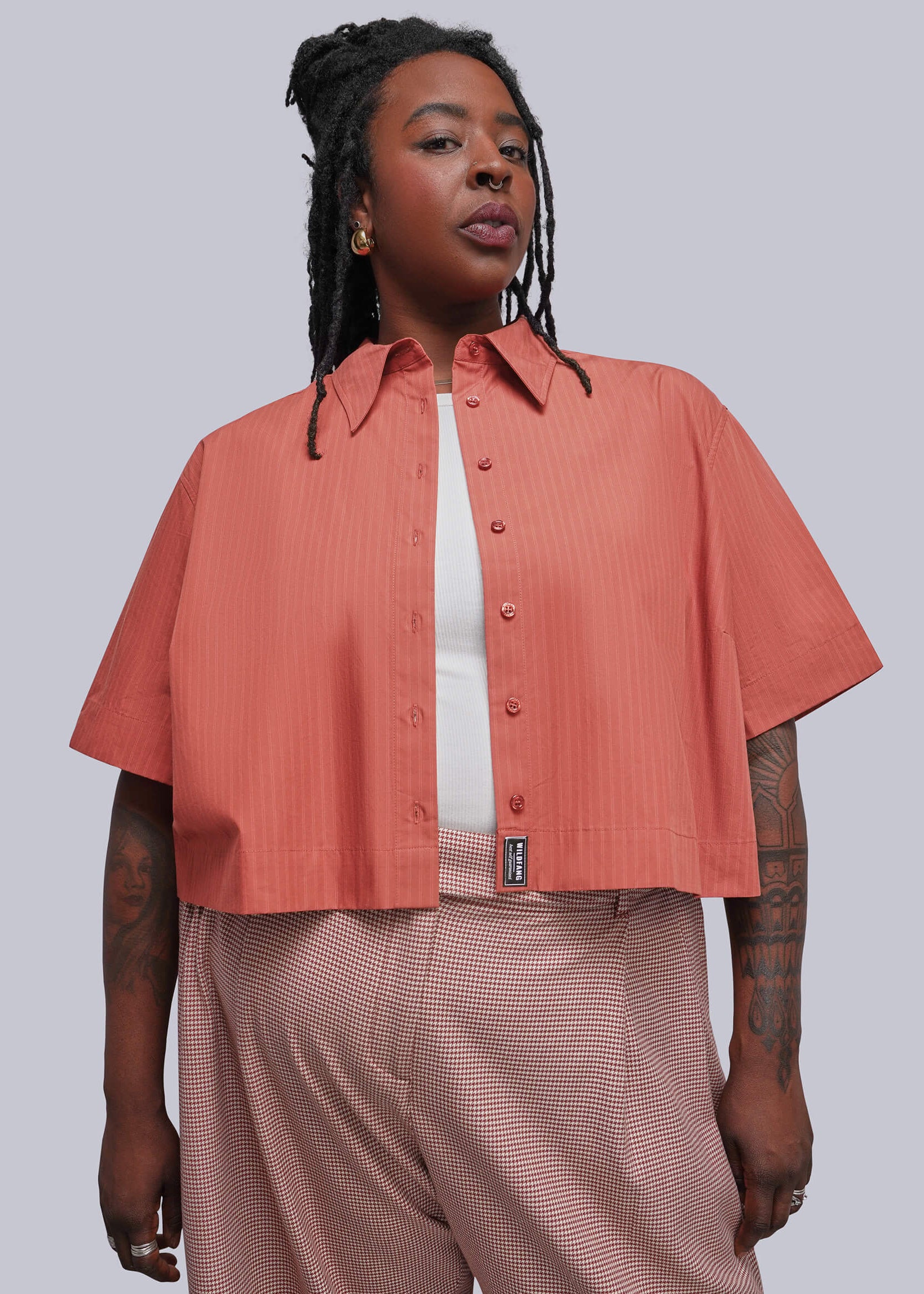 1X=A model wearing a short-sleeve cropped button-up shirt in a muted coral pink, styled over a white t-shirt and paired with herringbone patterned trousers, against a solid gray studio background.