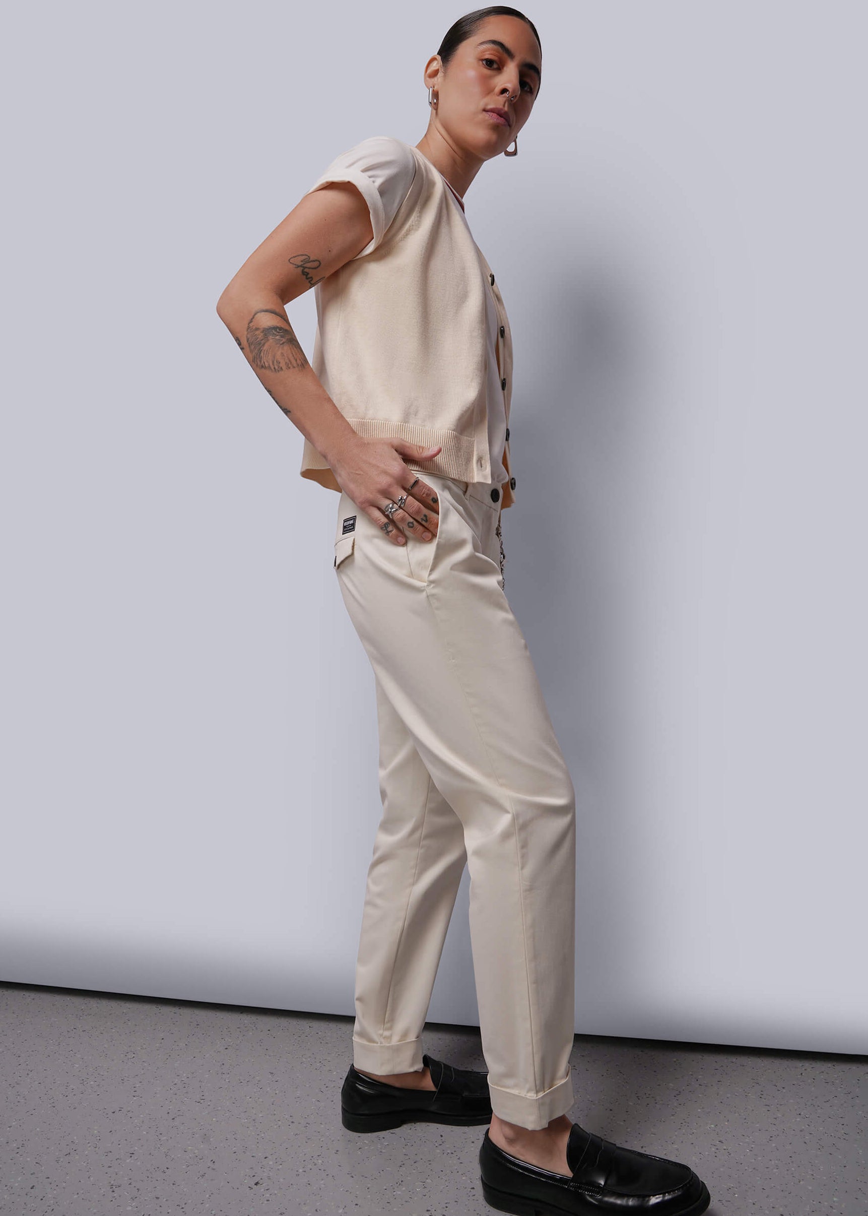 S=Person wearing a beige sweater vest and pants with black shoes against a plain background
