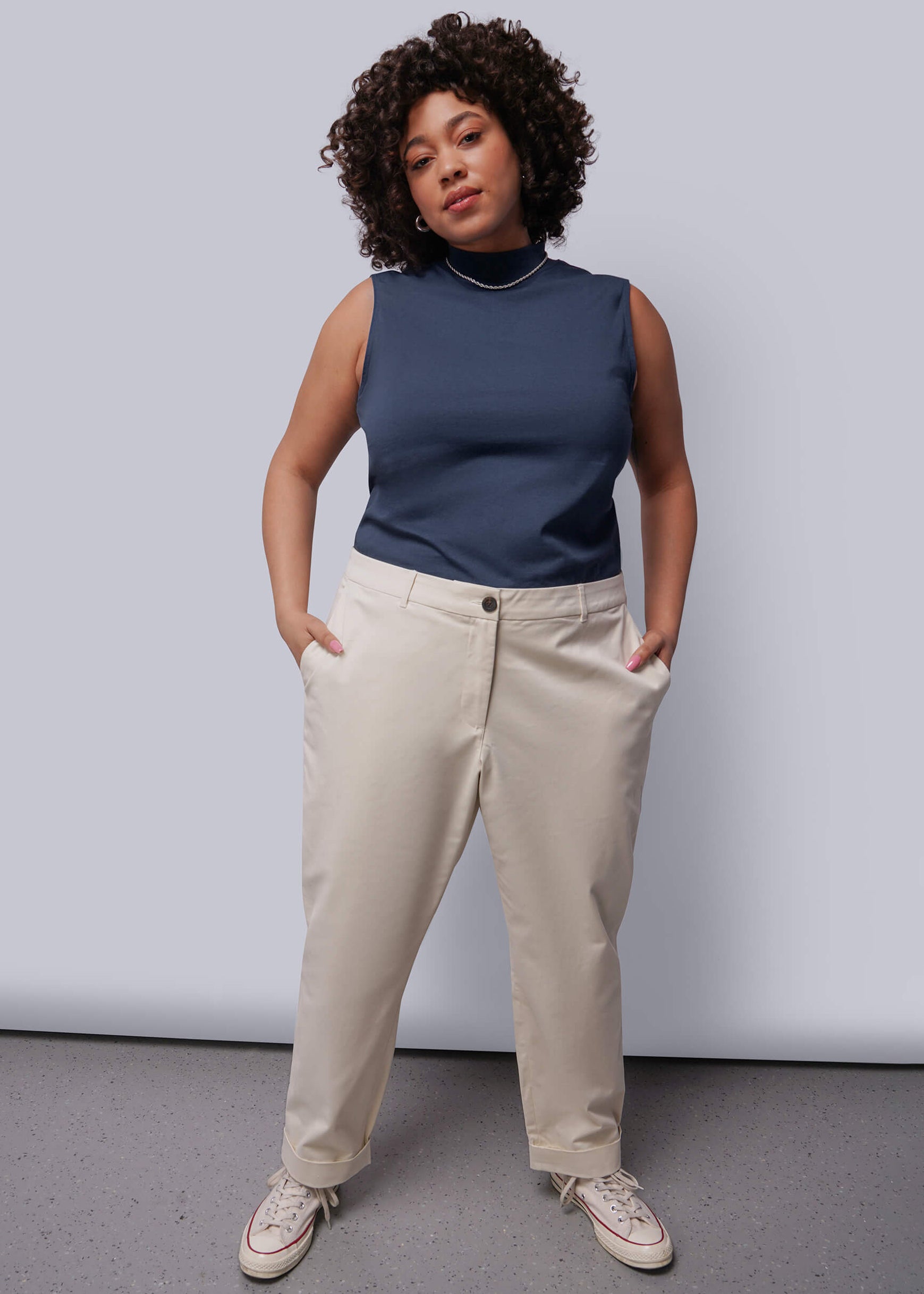 1X=Woman wearing a navy sleeveless top and beige pants against a plain background