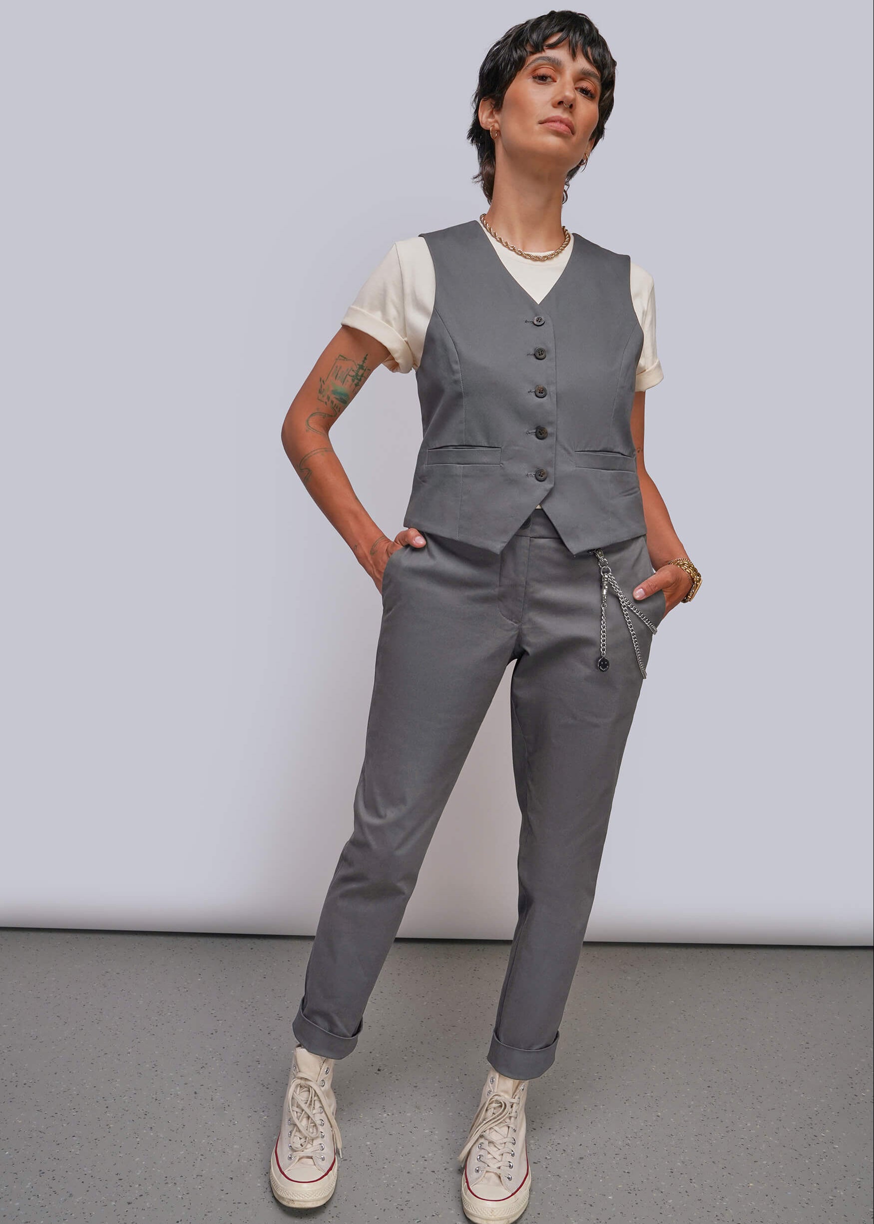 A model wearing a gray vest and matching gray trousers over a white t-shirt, styled with white sneakers, standing against a light gray studio background.