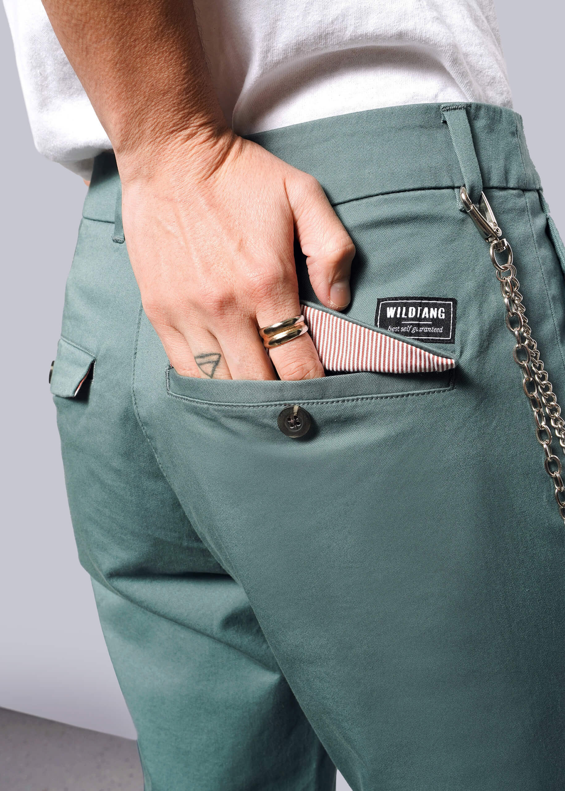 S=Wearing The Essential Trouser with a WILDIANG patch and relaxed mid rise fit, a person puts a striped wallet in their back pocket. Rings decorate their fingers, and a chain dangles from the classic trouser pocket.
