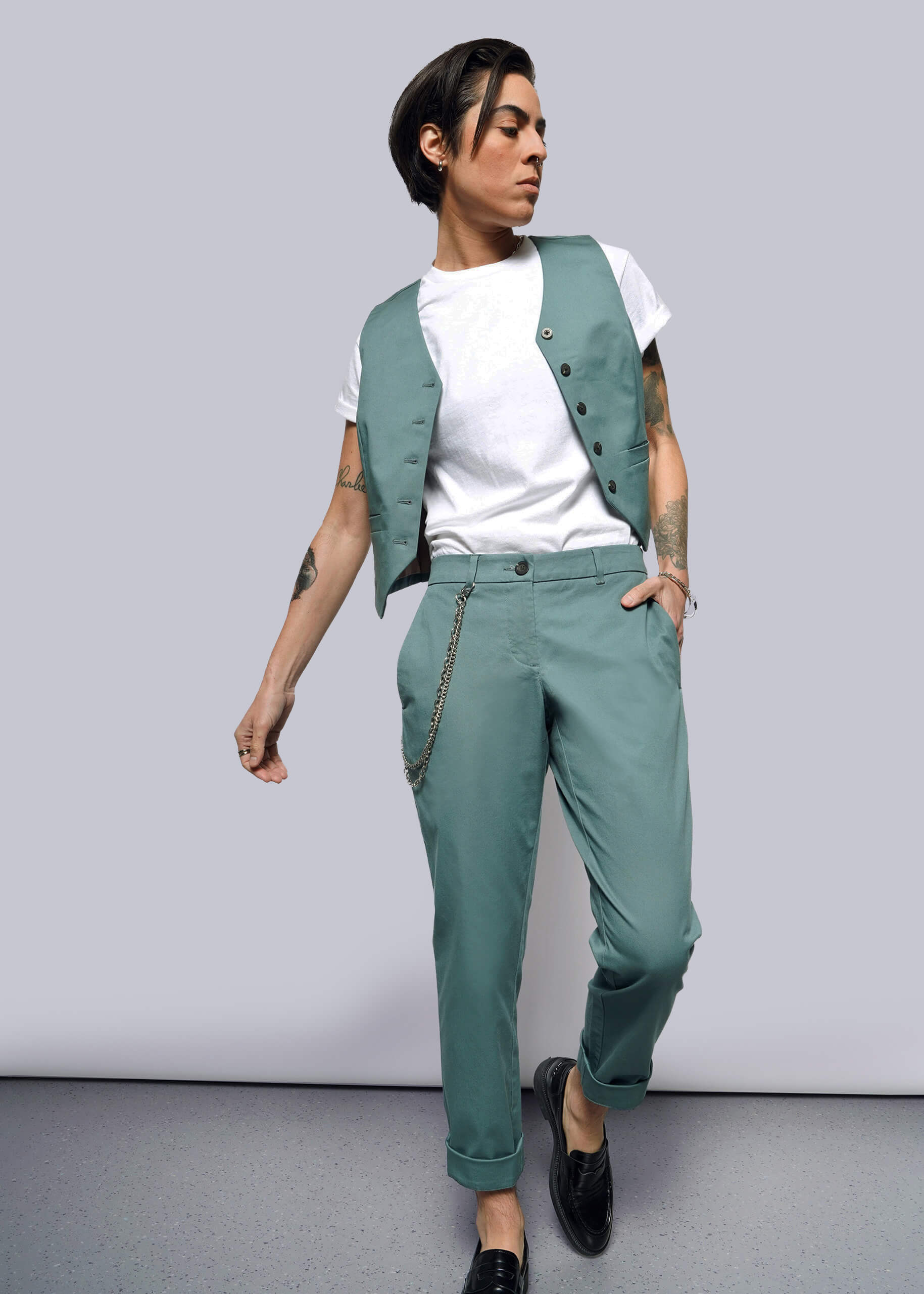 S=Person with short dark hair wears a white t-shirt, teal vest, and The Essential Trouser in a relaxed mid-rise fit with a chain accessory, standing against a plain white background and looking to the side.