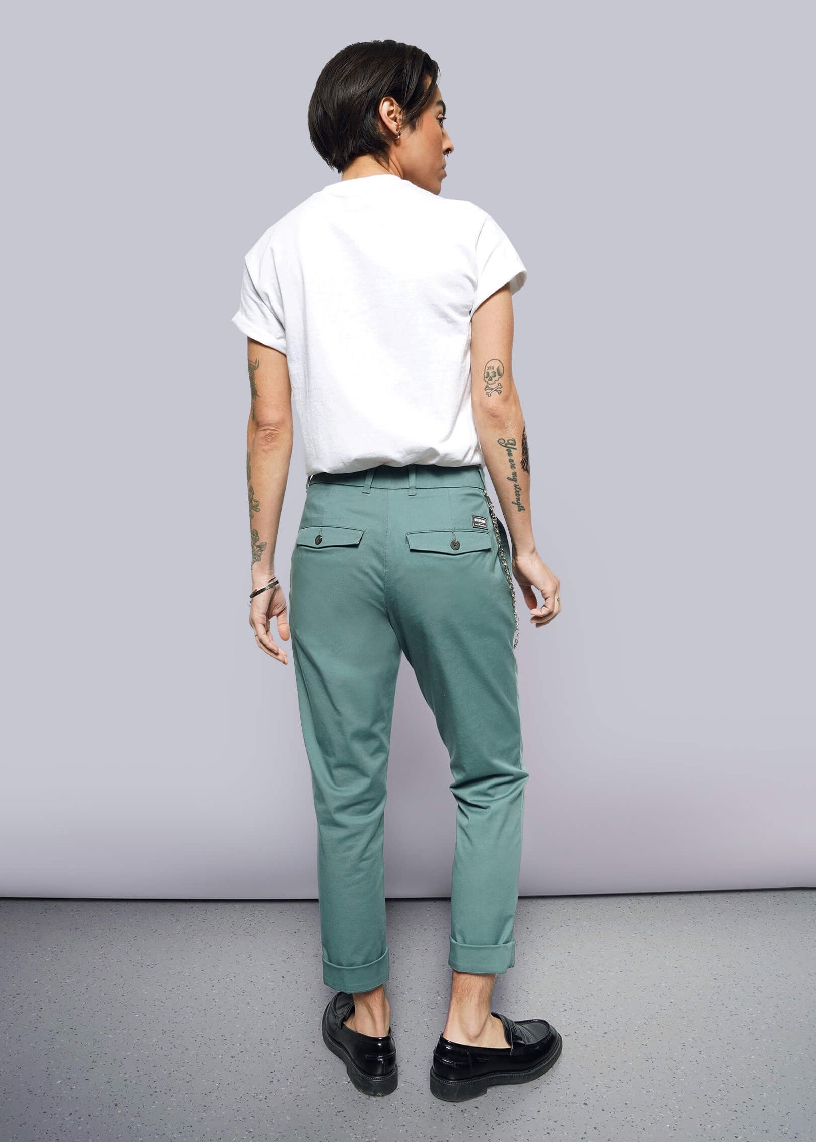 S= A person with short dark hair wearing a white rolled-sleeve tee, The Essential Trouser in teal with a relaxed mid-rise fit, and black loafers stands facing away. Tattoos are visible on both arms. The background is plain and well-lit.