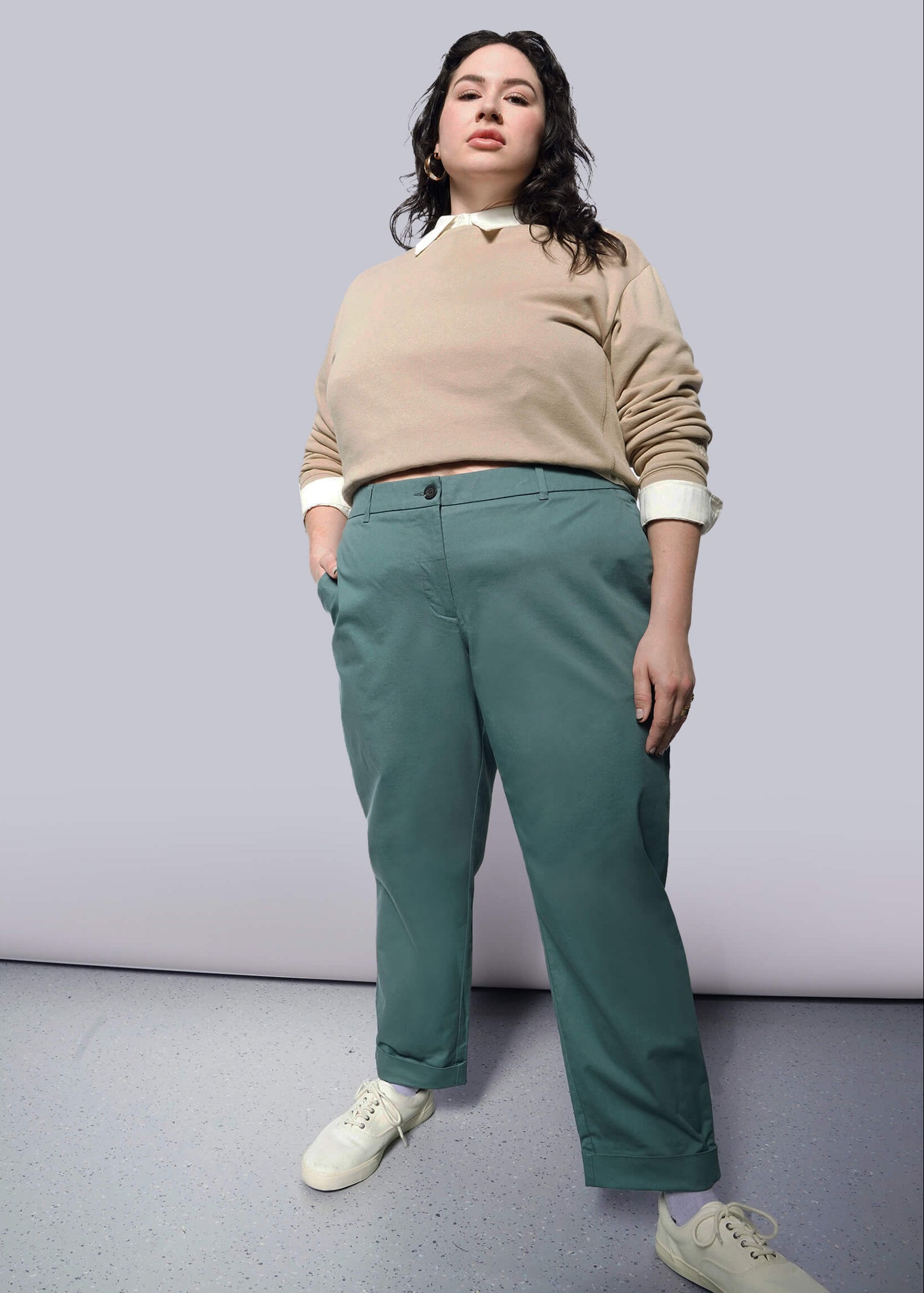 1X= A person with long dark hair stands confidently with one hand in their pocket, wearing a beige sweater, The Essential Trouser in teal with a relaxed mid rise fit, and white sneakers against a plain light background.