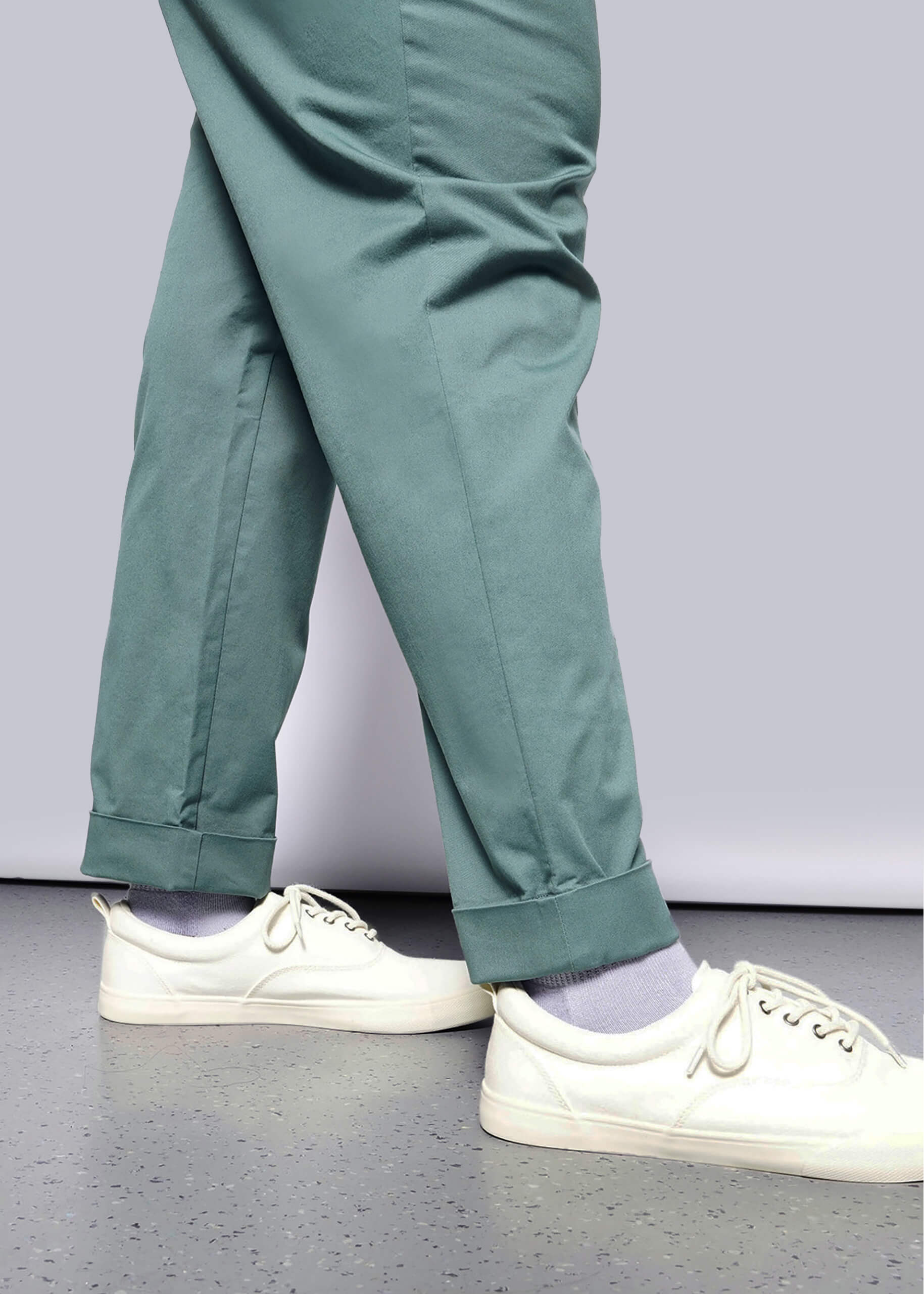 1X=A person wearing The Essential Trouser in teal with a relaxed mid-rise fit, paired with light purple socks and white sneakers, walks on a light floor. Only their legs and feet are visible against a white background.