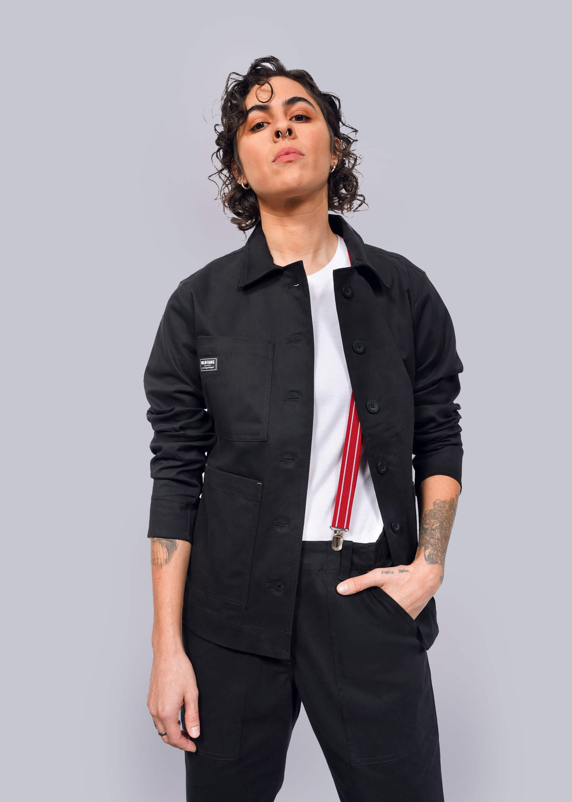Wearing The Essential Utility Jacket and comfortable stretch pants, a person with short curly hair pairs their look with a white shirt and red suspenders, standing confidently against a white background.