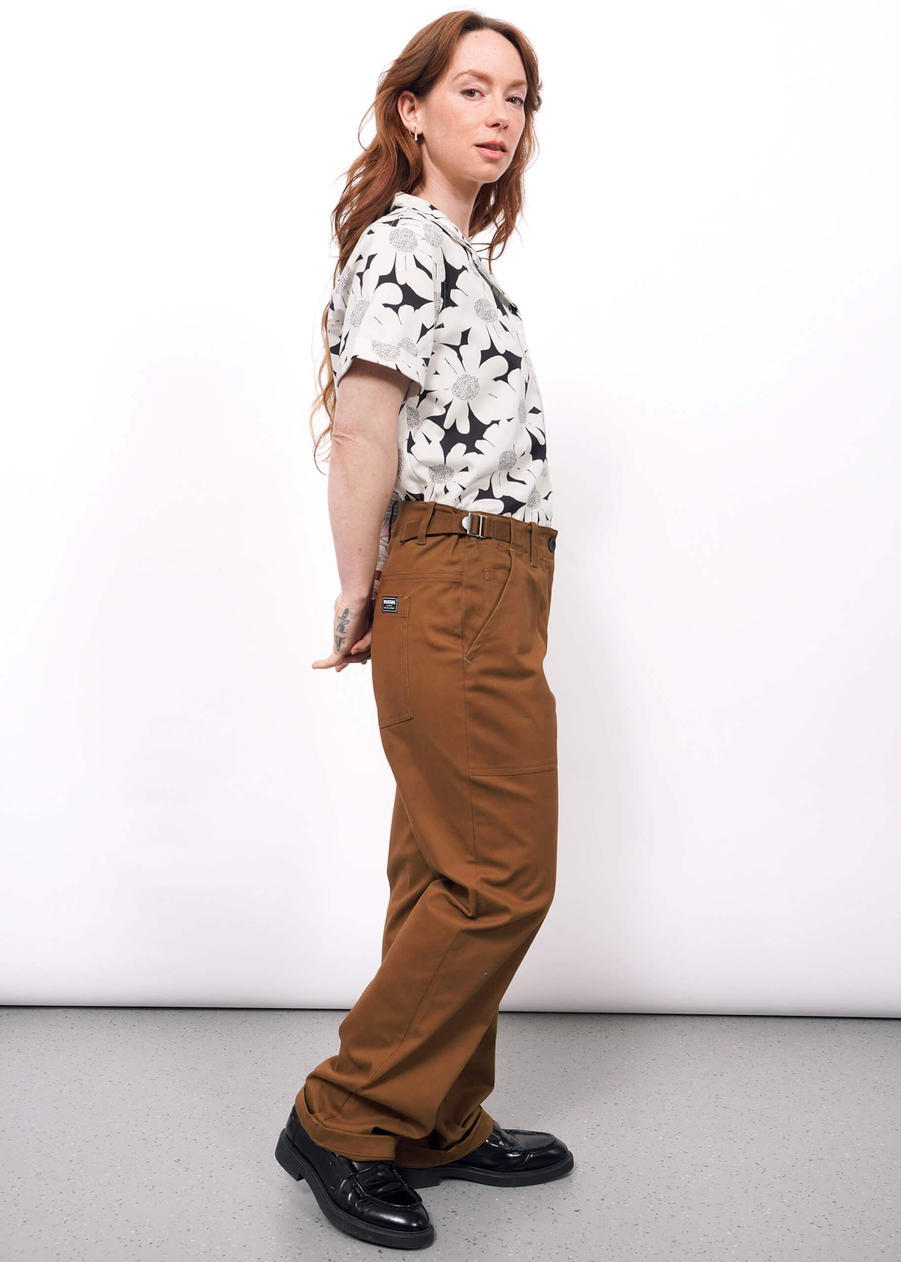 S=A person with long, wavy hair stands sideways against a white wall, wearing The Essential Utility Pant—high-waisted brown pants with an adjustable waist—paired with a floral shirt and black shoes, hands clasped behind their back.
