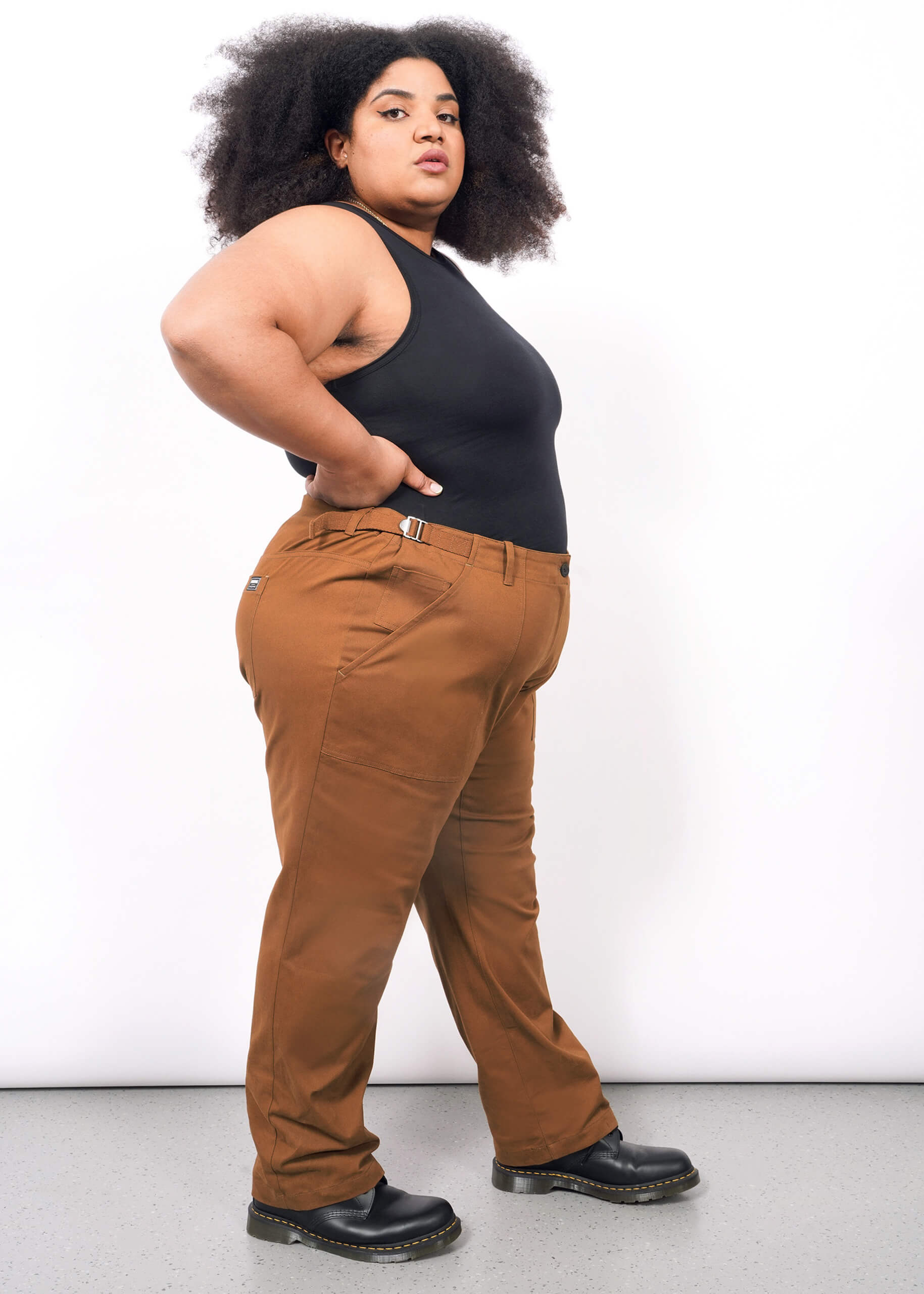 1X=A woman with curly hair models The Essential Utility Pant—rust-colored with roomy pockets and side buckles—paired with a sleeveless black top and black shoes, standing sideways against a white background.