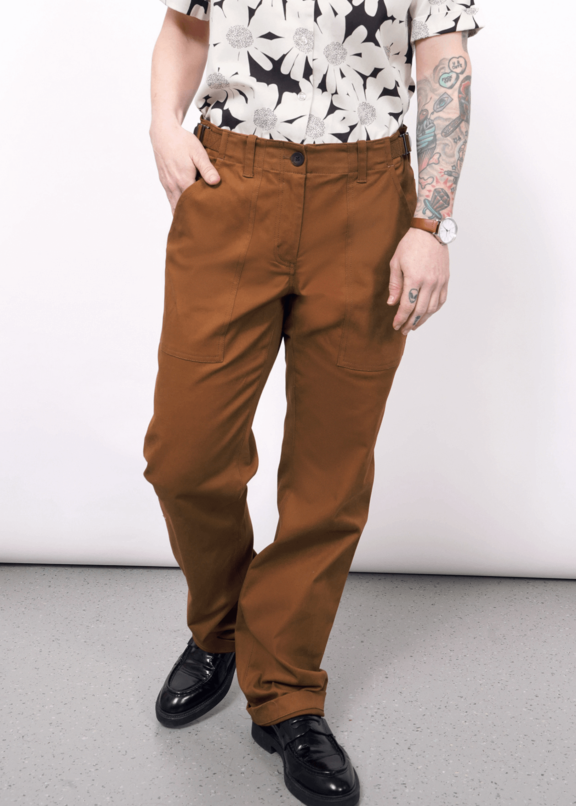 S=A person stands against a light background wearing The Essential Utility Pant in rust, featuring spacious pockets, paired with black loafers and a black-and-white floral shirt. One hand is in a pocket; the other arm with tattoos rests by their side.
