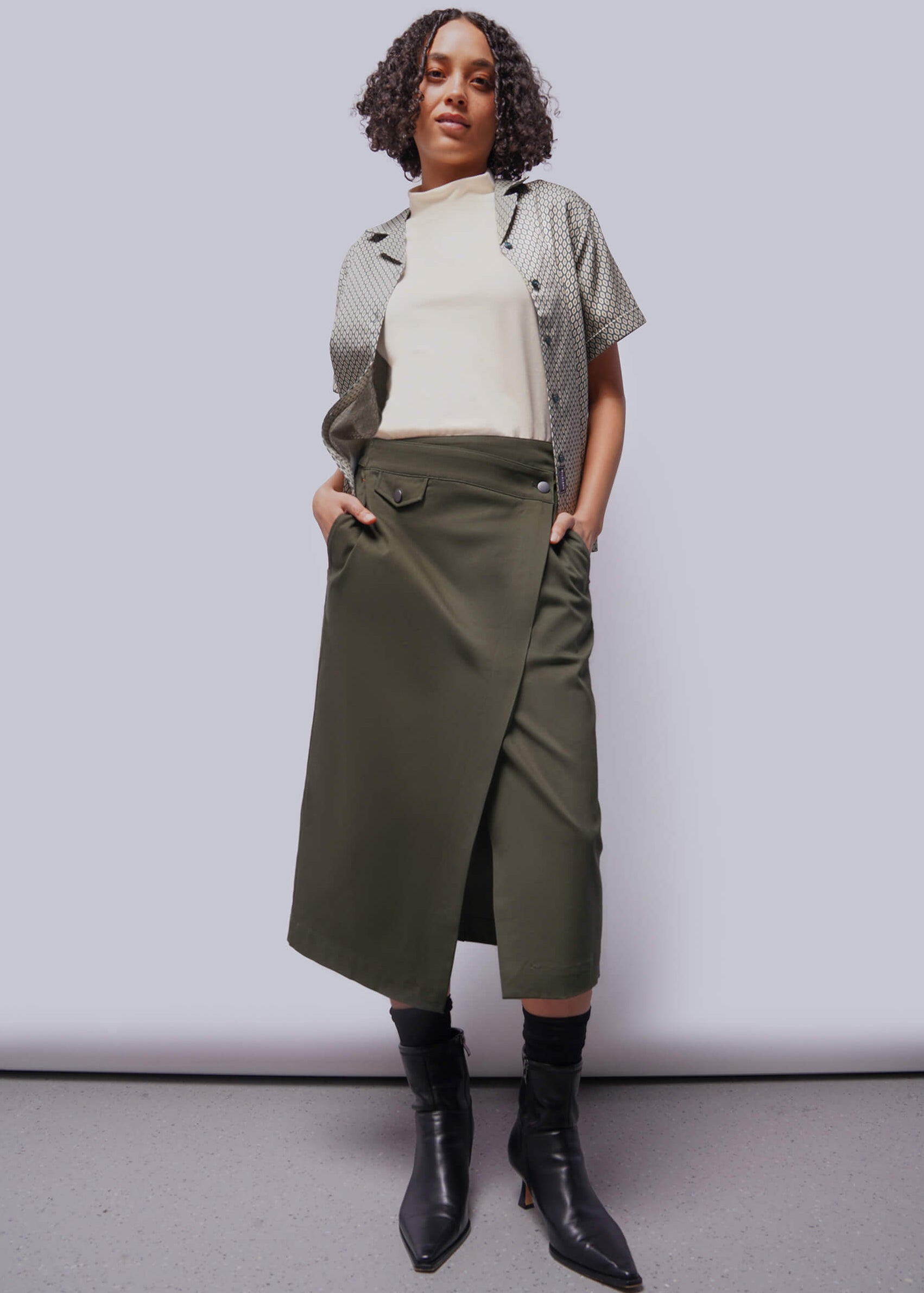 Person wearing a white shirt, patterned cardigan, green skirt, and black boots against a plain background