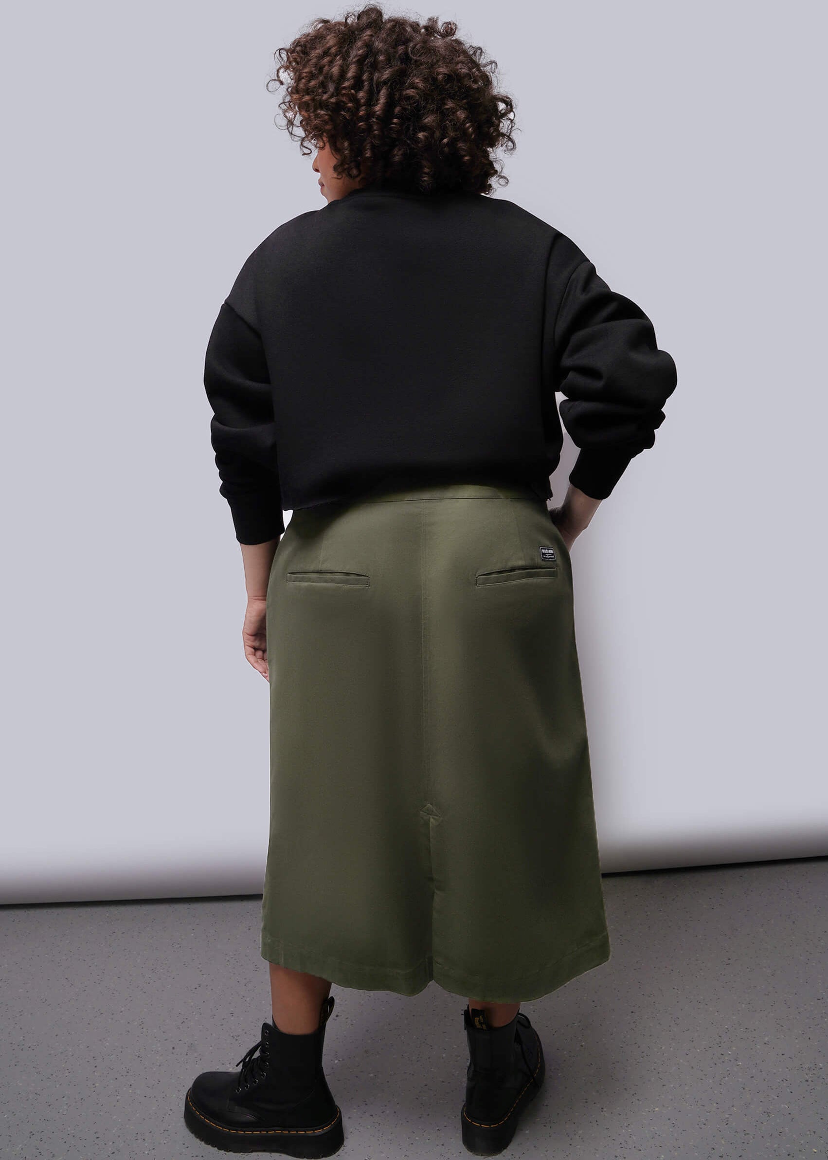 1X=Person wearing a black sweatshirt and green skirt against a white background