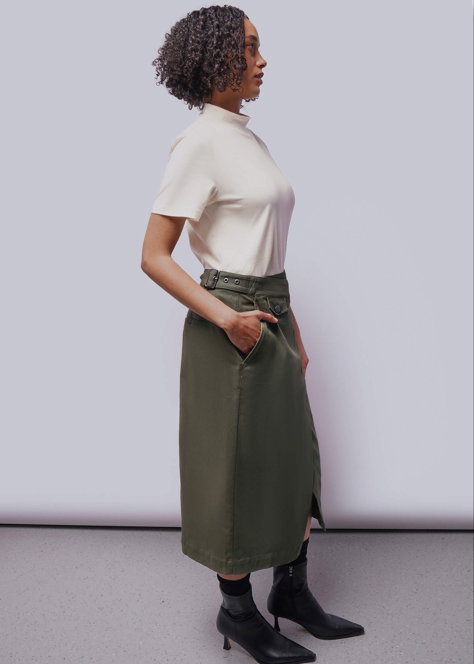 S=Side view of person wearing cream colored shirt and green skirt with black boots