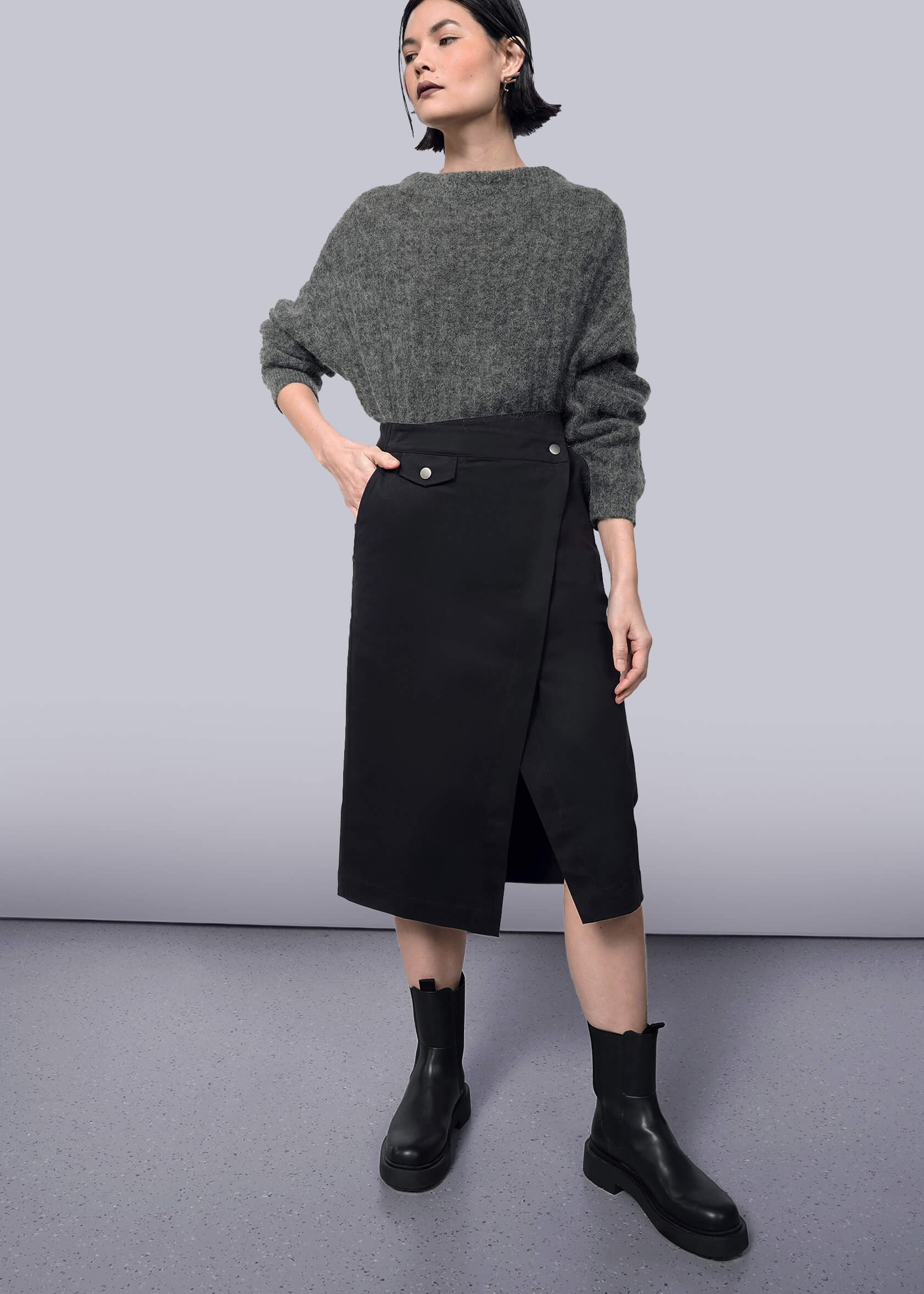  A woman poses against a white backdrop in a gray sweater and The Essential Utility Wrap Skirt—black, midi length, with buttons and an asymmetric waist. She pairs the look with black ankle boots and styles her short dark hair confidently.