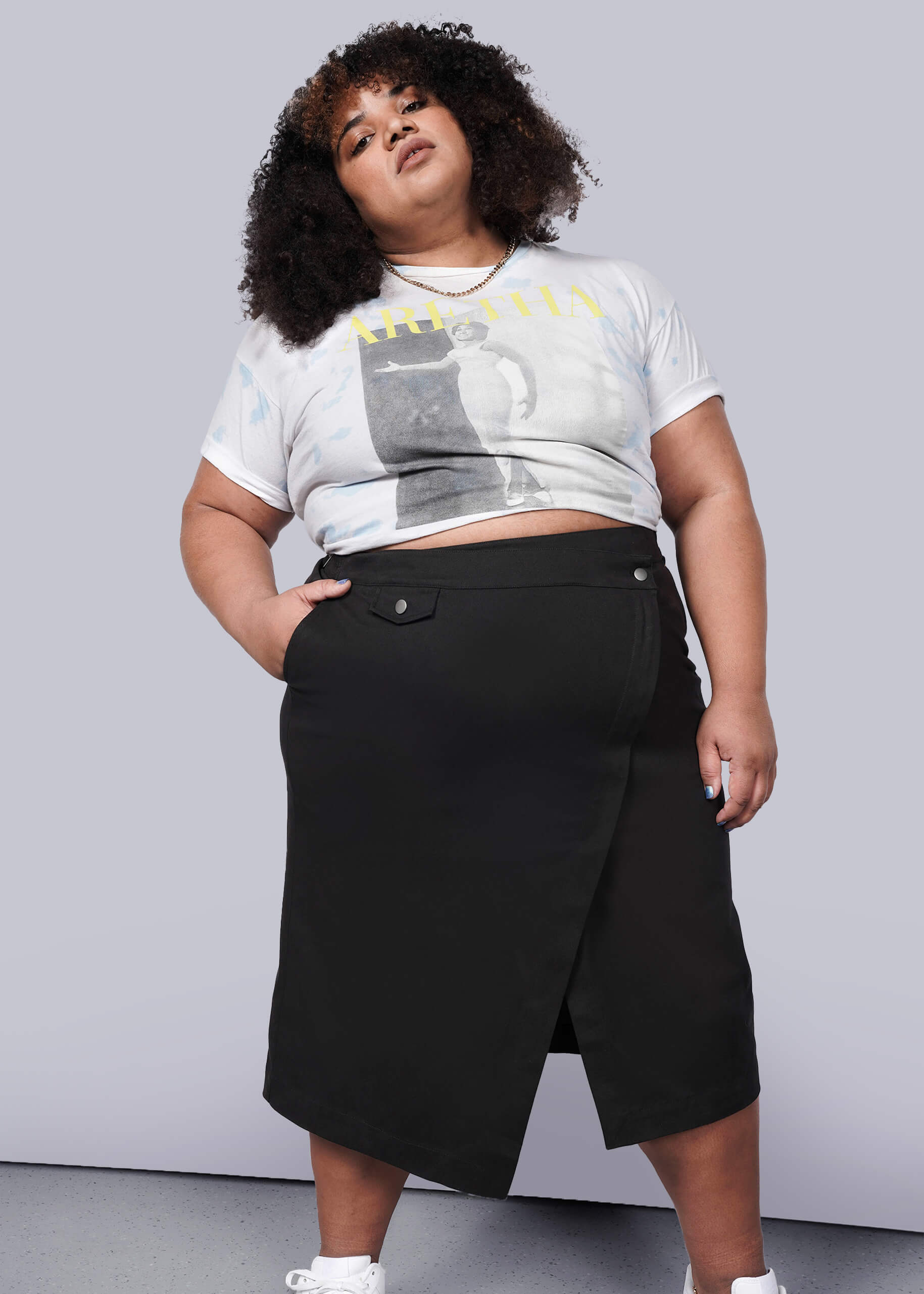 1X= A person with curly hair wears a printed crop top, The Essential Utility Wrap Skirt in black with an asymmetrical waist detail, and white sneakers, standing against a plain white wall and looking confidently at the camera.