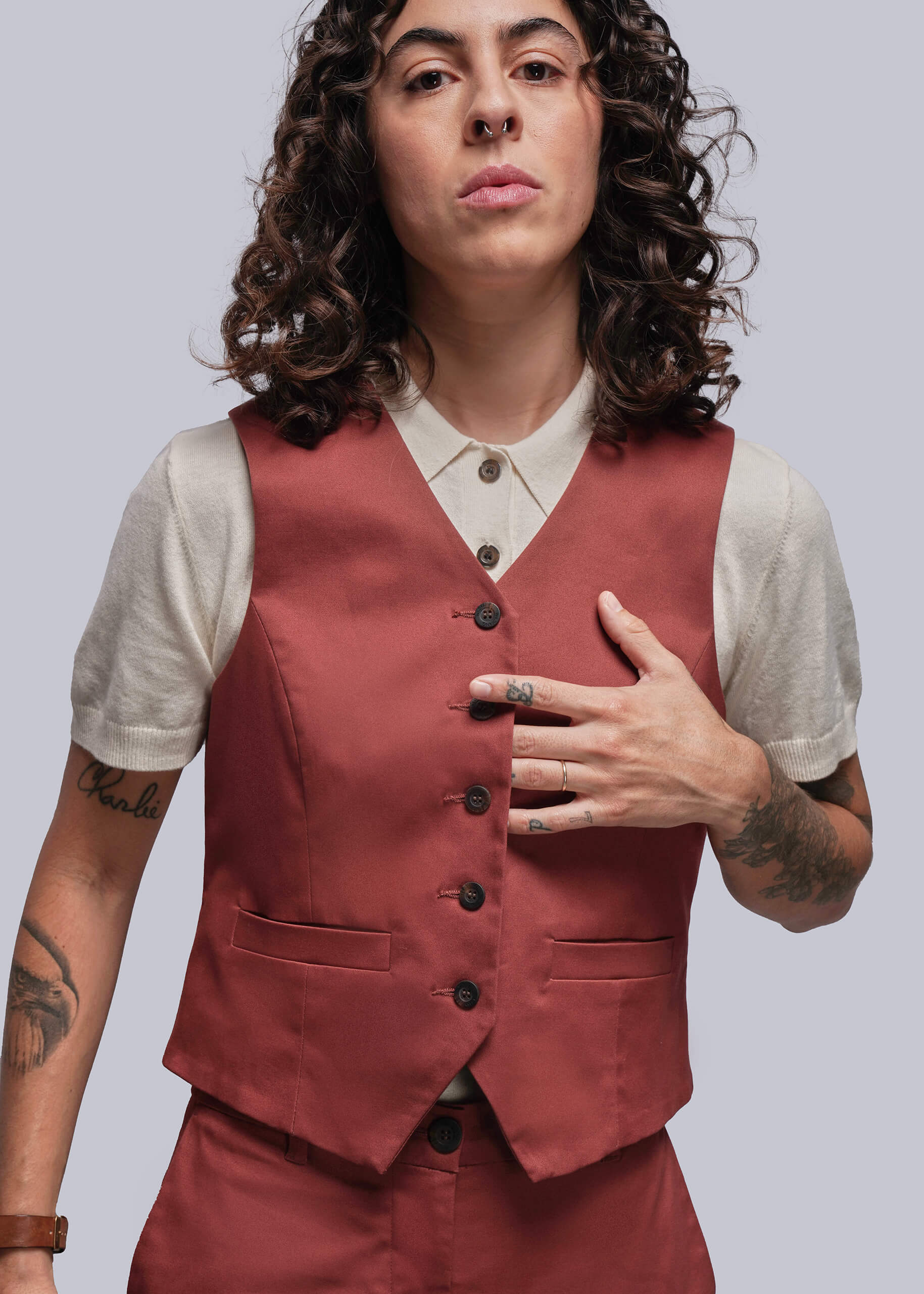 Person wearing a rust colored vest over a white sweater polo with tattoos on arms, standing against a plain background
