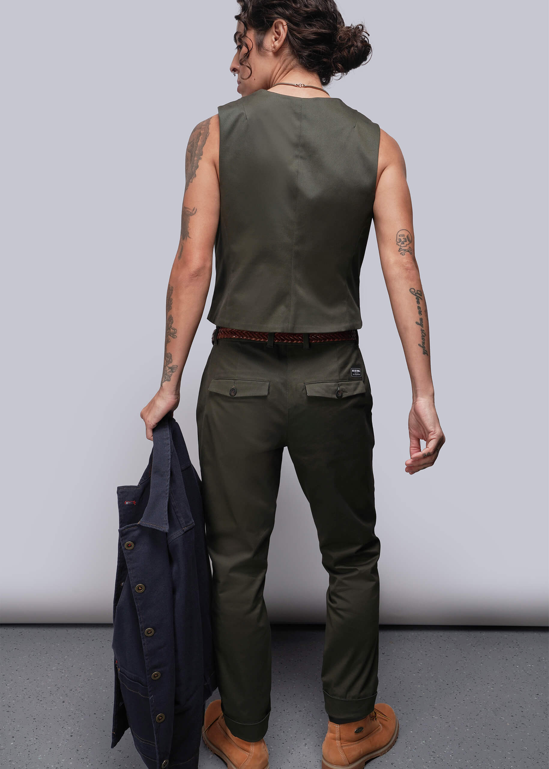 S=Person wearing a green sleeveless vest and pants holding a jacket against a plain background