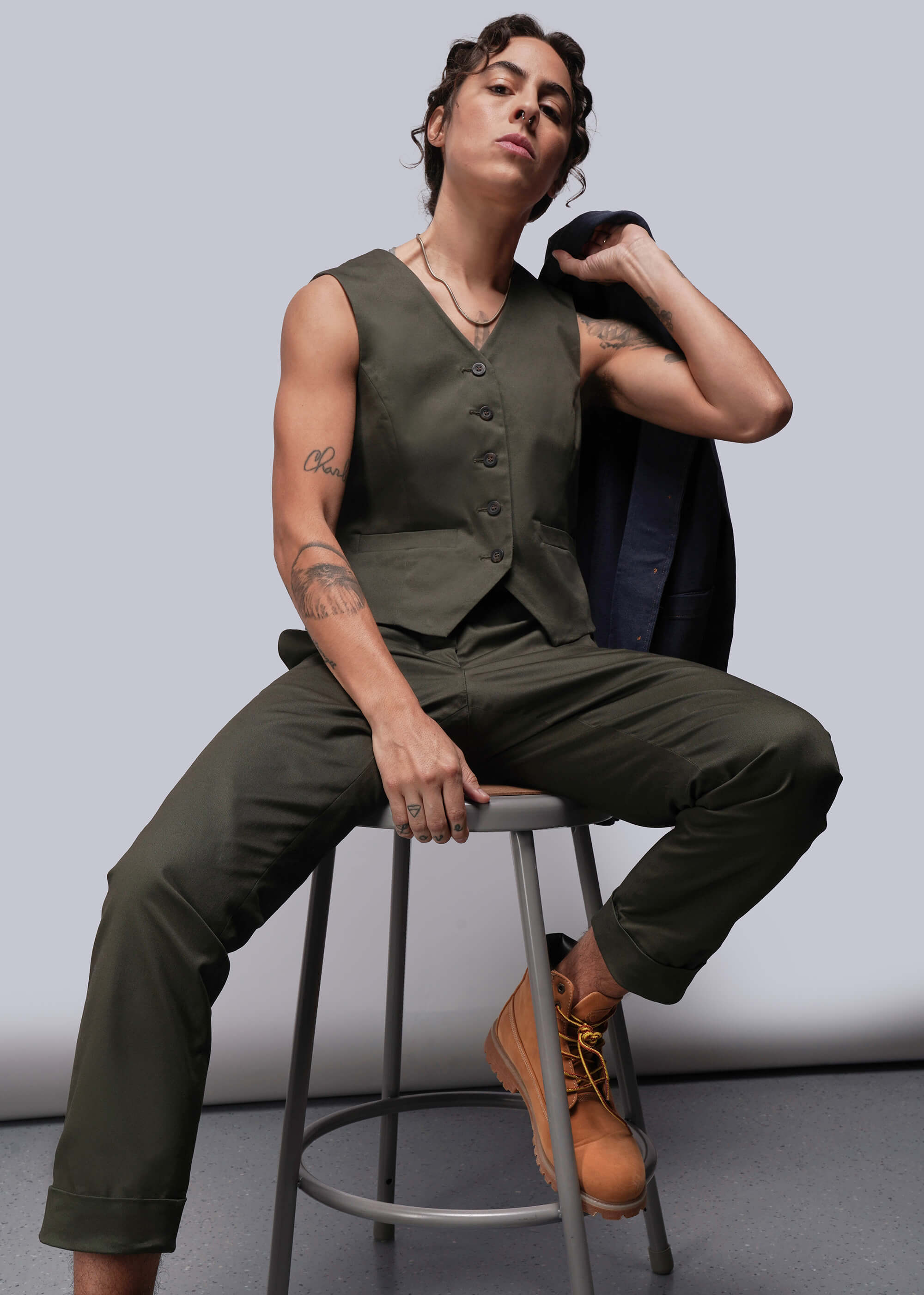Person wearing a green vest and pants sitting on a stool against a plain background