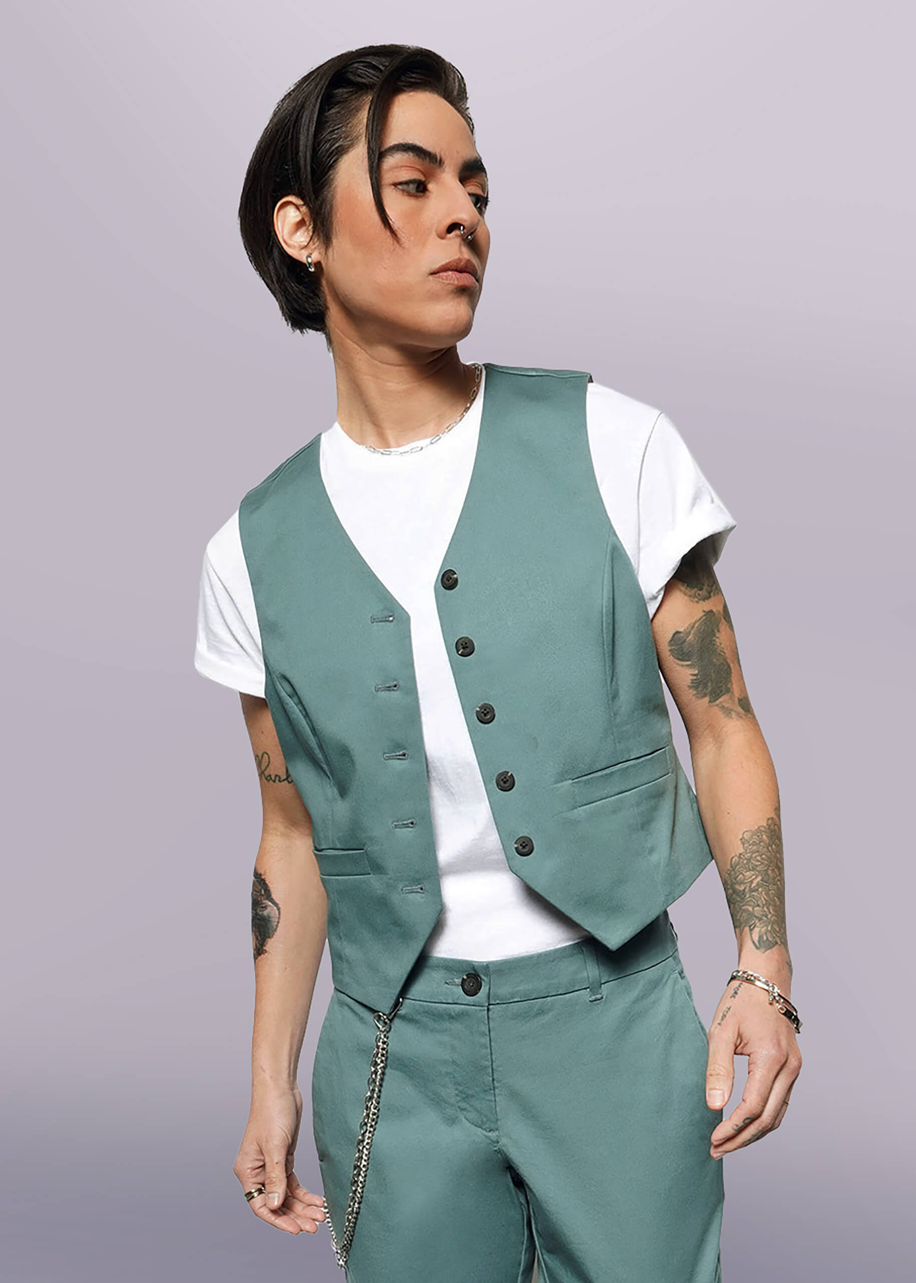 A person with short dark hair wears The Essential Vest and matching stretch cotton pants over a white t-shirt, accessorized with bracelets and a chain, standing against a plain white background.