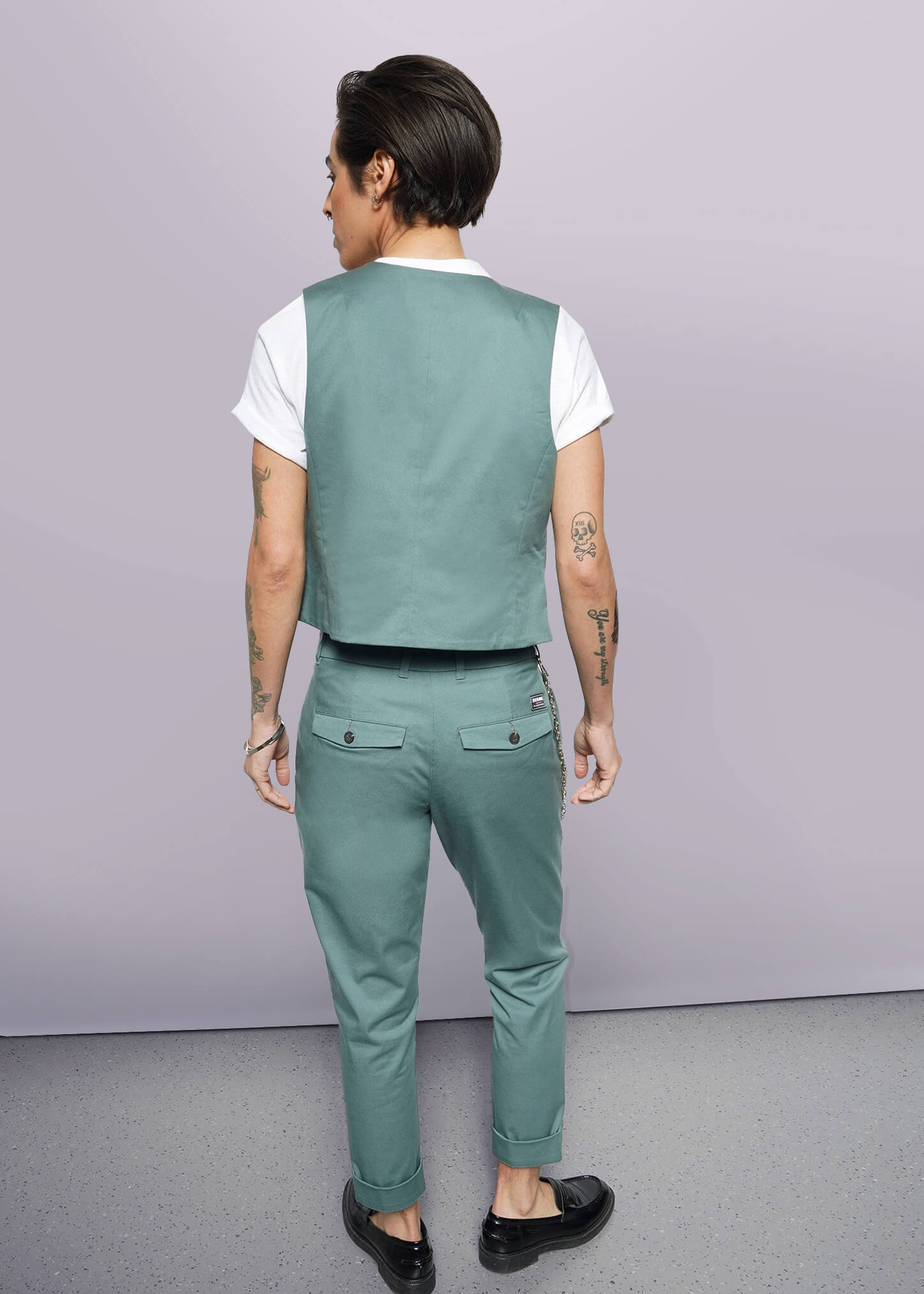 A person with short dark hair, wearing The Essential Vest—tailored fit, stretch cotton, 5-button closure—with matching pants, white T-shirt, and black shoes stands facing away from the camera, displaying arm tattoos against a plain white background.