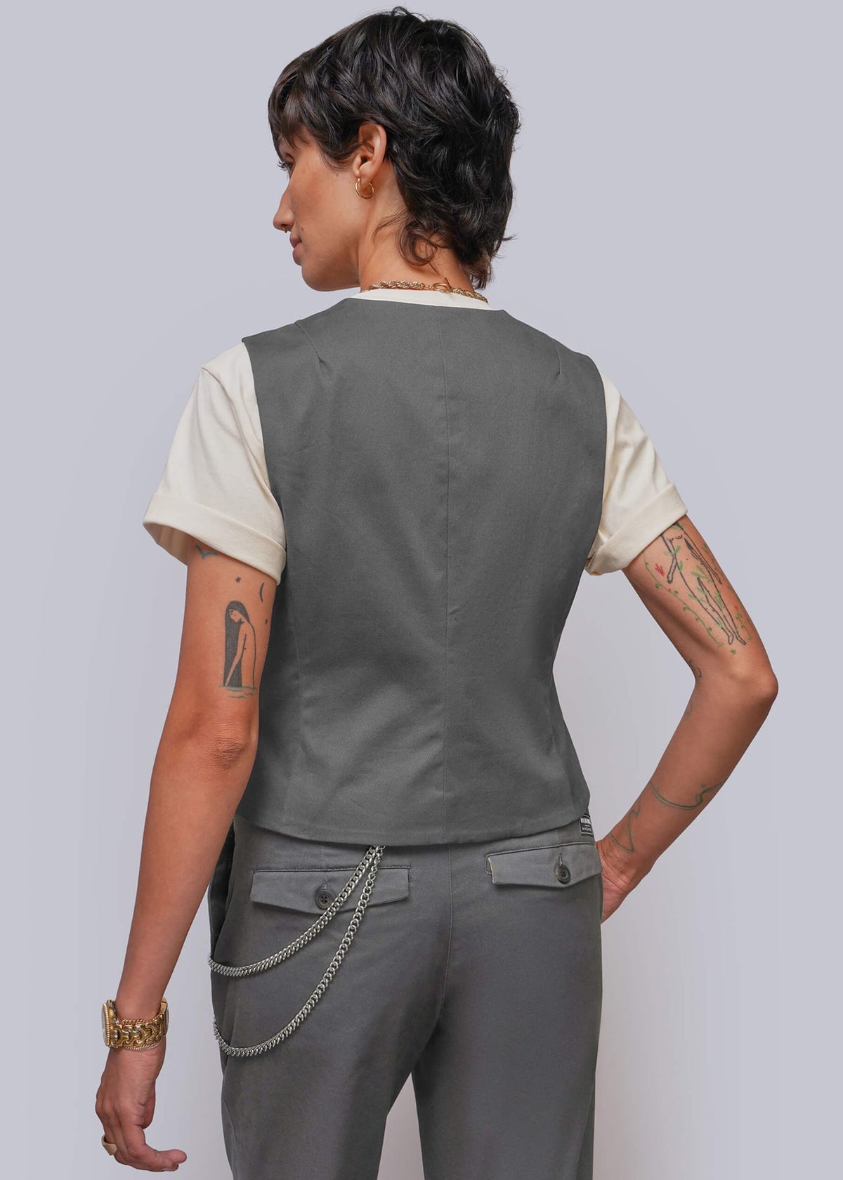S=A model shown from the back wearing a dark gray sleeveless vest over a cream t-shirt and matching gray trousers with a silver chain detail at the back waist.