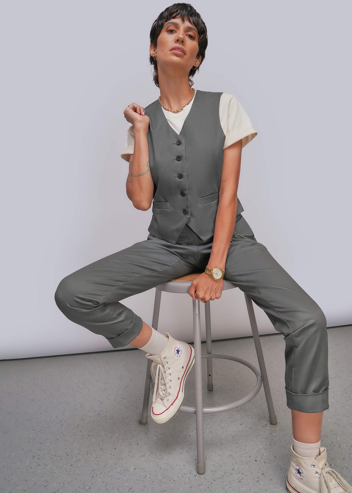 S=A model sits on a metal stool wearing a gray button-front vest, matching gray cropped trousers, a white short-sleeve top, and white high-top sneakers against a light gray studio background.