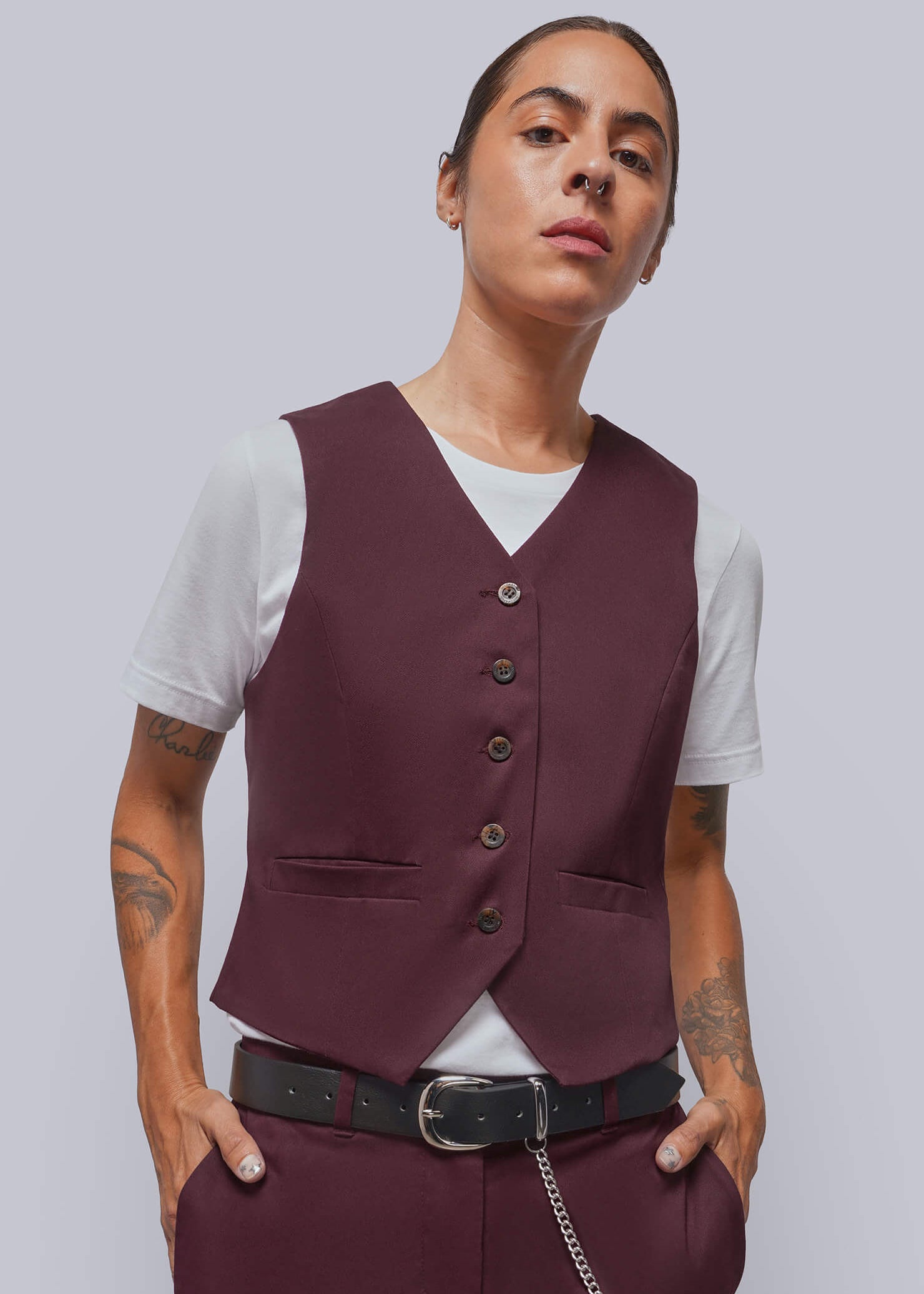 A model wearing a structured burgundy vest over a white t-shirt, paired with matching burgundy trousers, a black belt, and a silver chain, set against a solid light gray background.